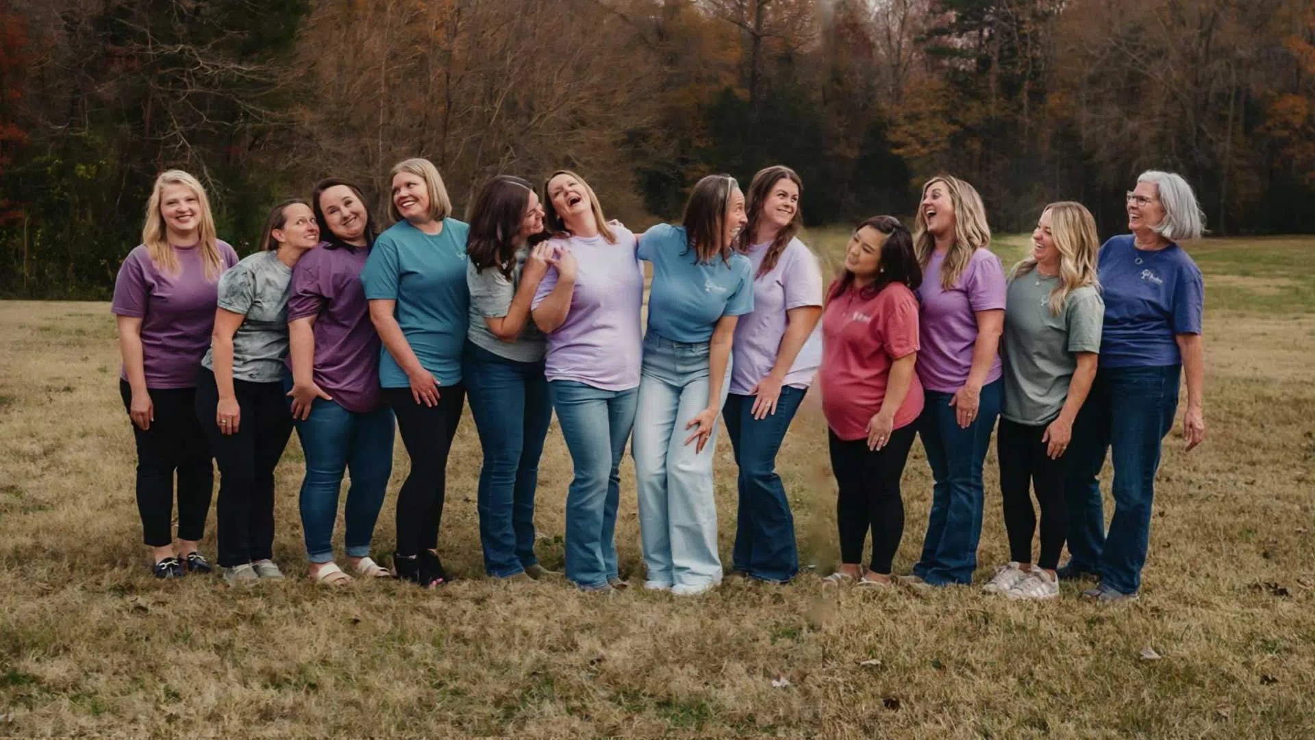 Group of women standing together laughing outdoors in casual colorful shirts and jeans on grassy field