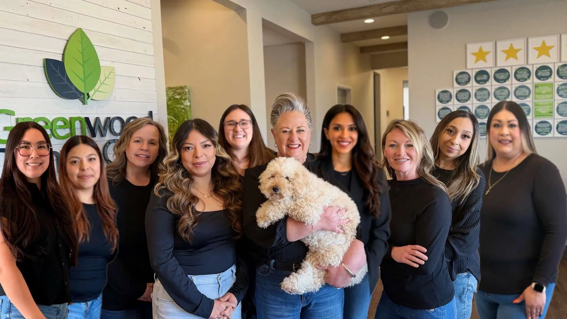 Group of smiling women wearing black tops posing indoors with a fluffy white dog in front of a Greenwood sign.