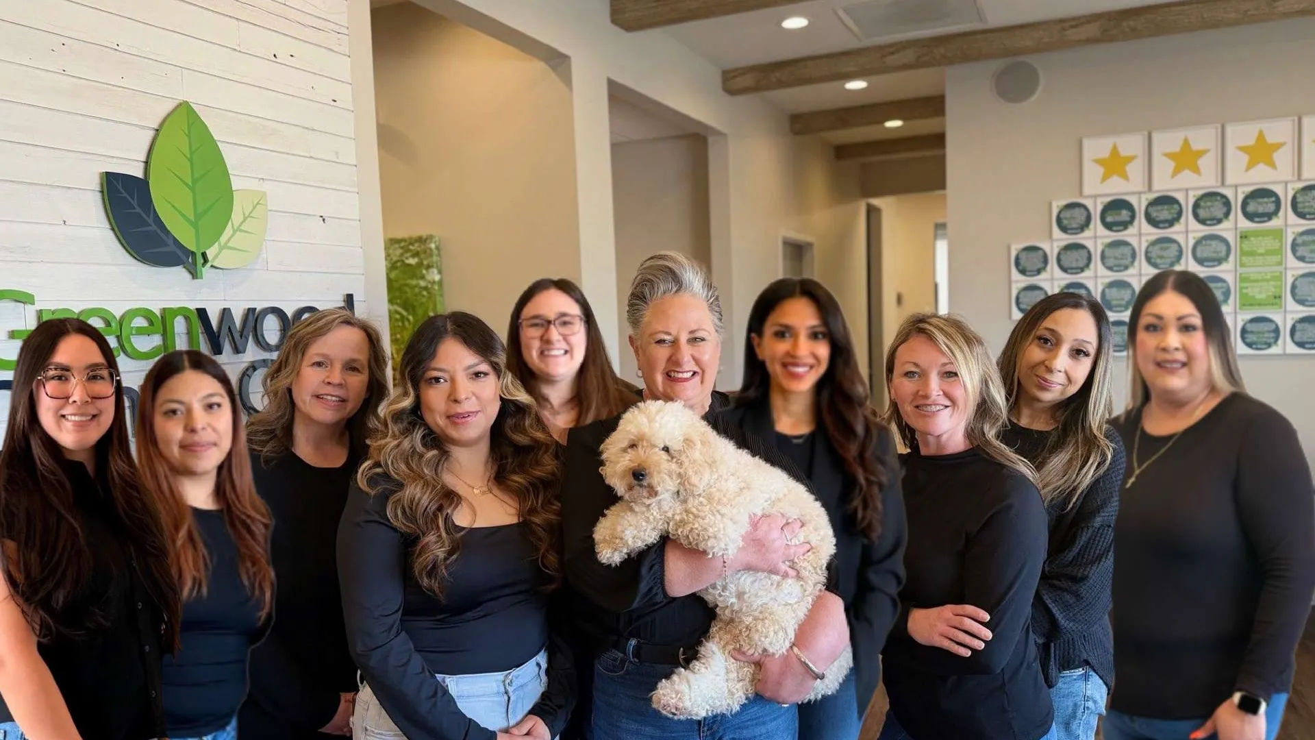 Group of smiling women wearing black tops posing indoors with a fluffy white dog in front of a Greenwood sign.