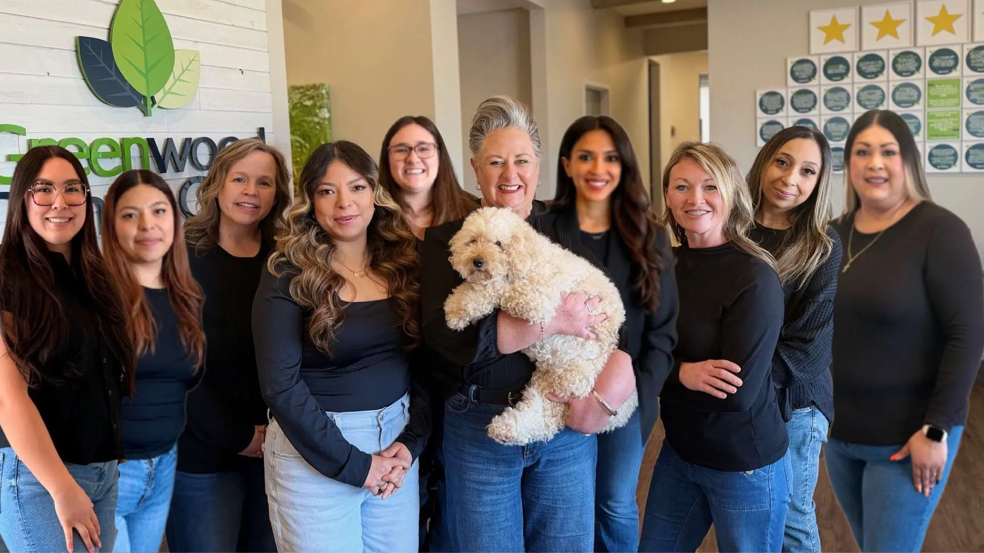 Group of smiling women wearing black tops posing indoors with a fluffy white dog in front of a Greenwood sign.