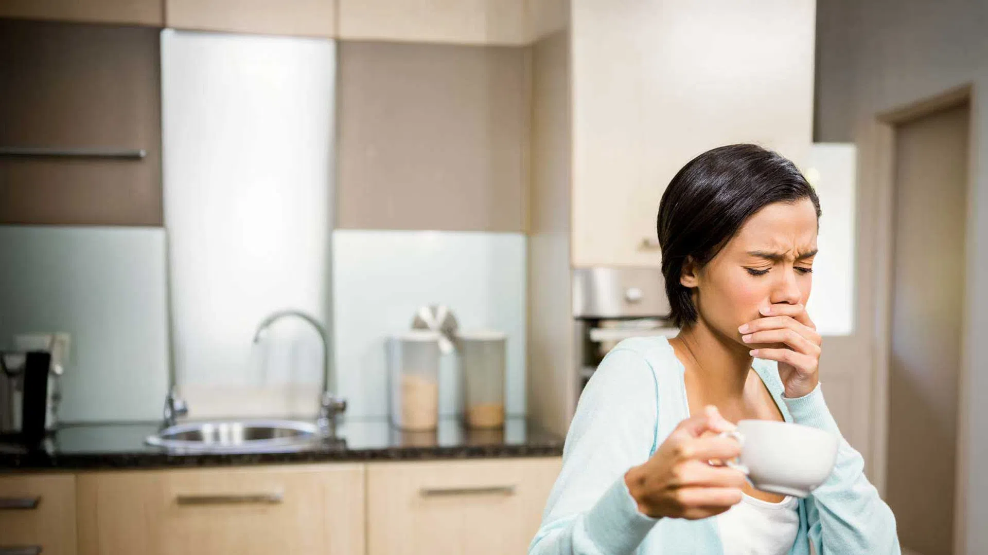 Woman holding a cup, feeling nauseous in a modern kitchen with wooden cabinets and a coffee maker.