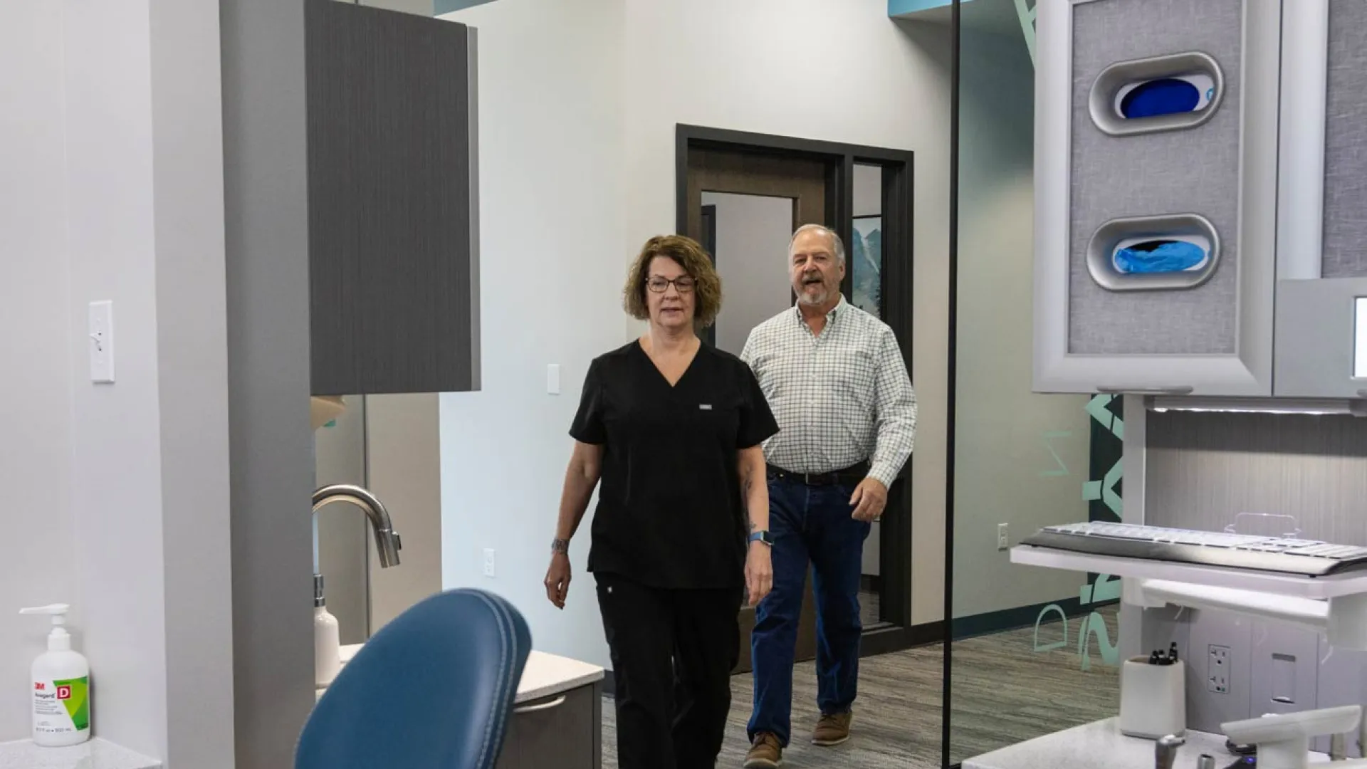 Dental office interior with a female dental professional and male patient walking through modern treatment area.