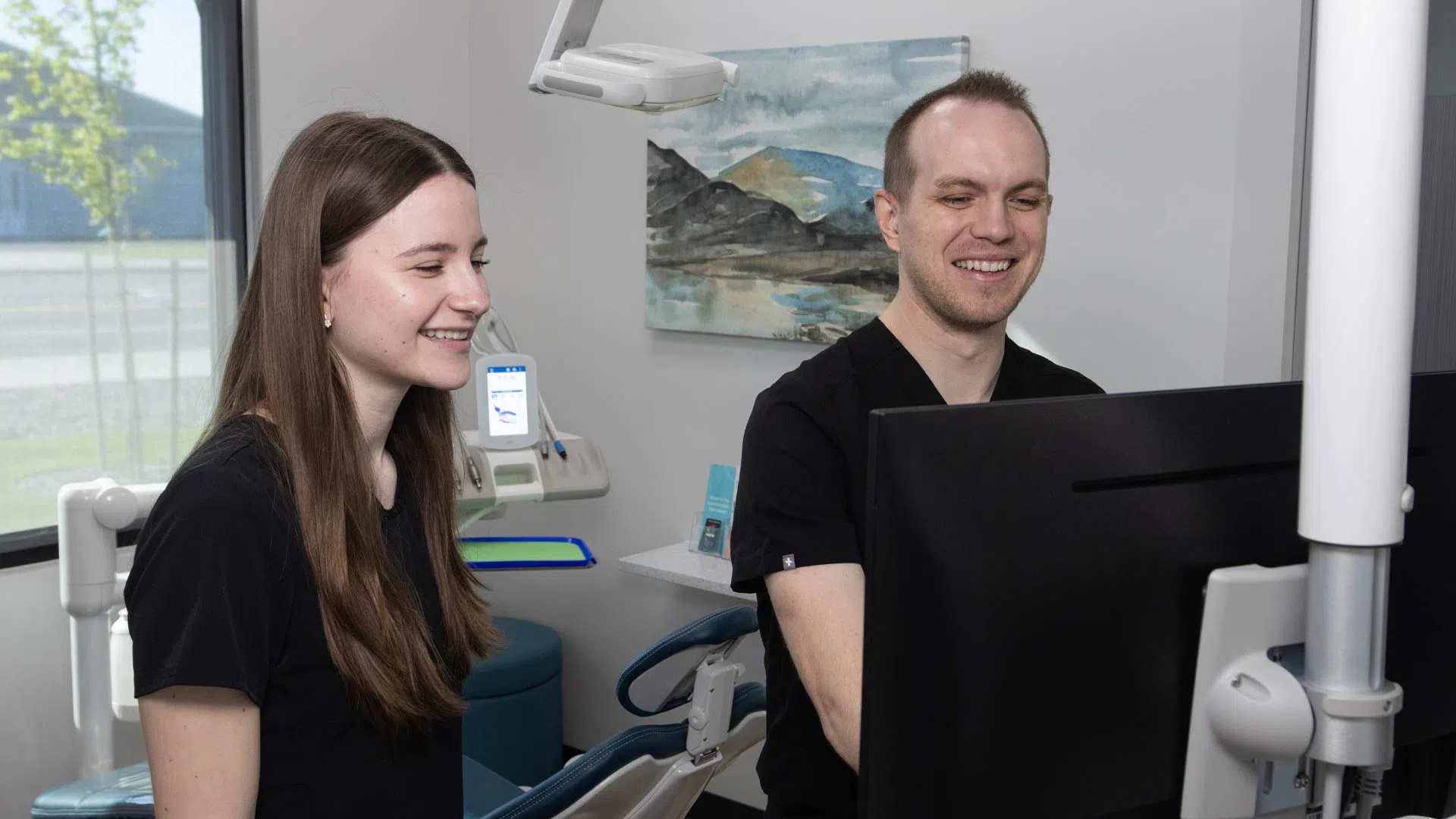 Two dental professionals smiling while reviewing information on a computer screen in a dental office.