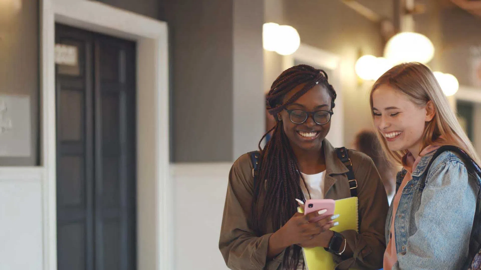 Two diverse young women smiling and looking at a smartphone in a brightly lit hallway.