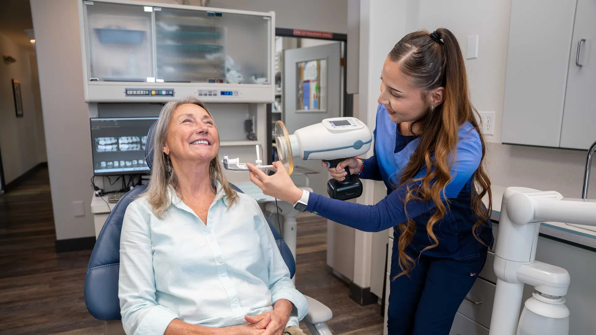 Dental hygienist using handheld imaging device to examine smiling senior woman in dental office chair