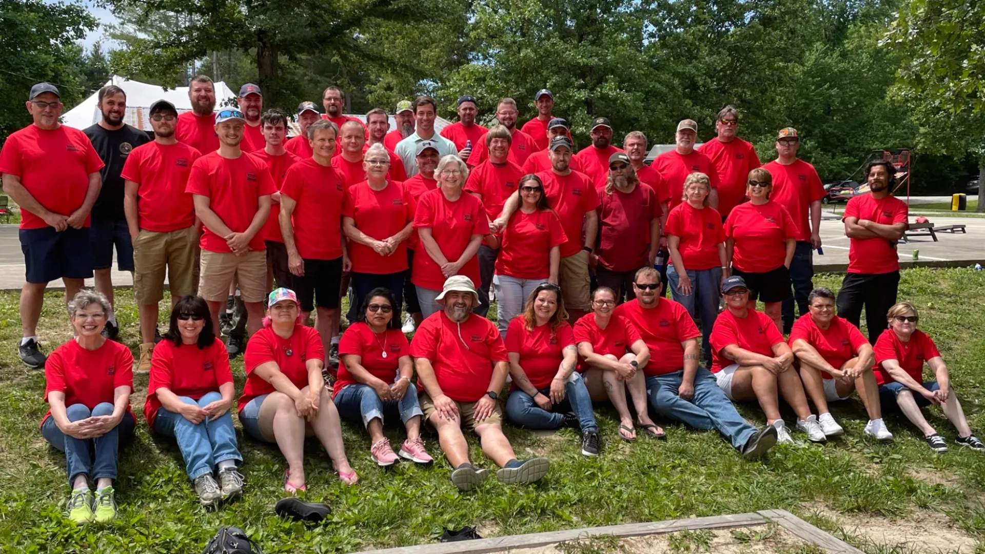 Large group of people wearing matching red shirts posing outdoors on grass under trees on a sunny day