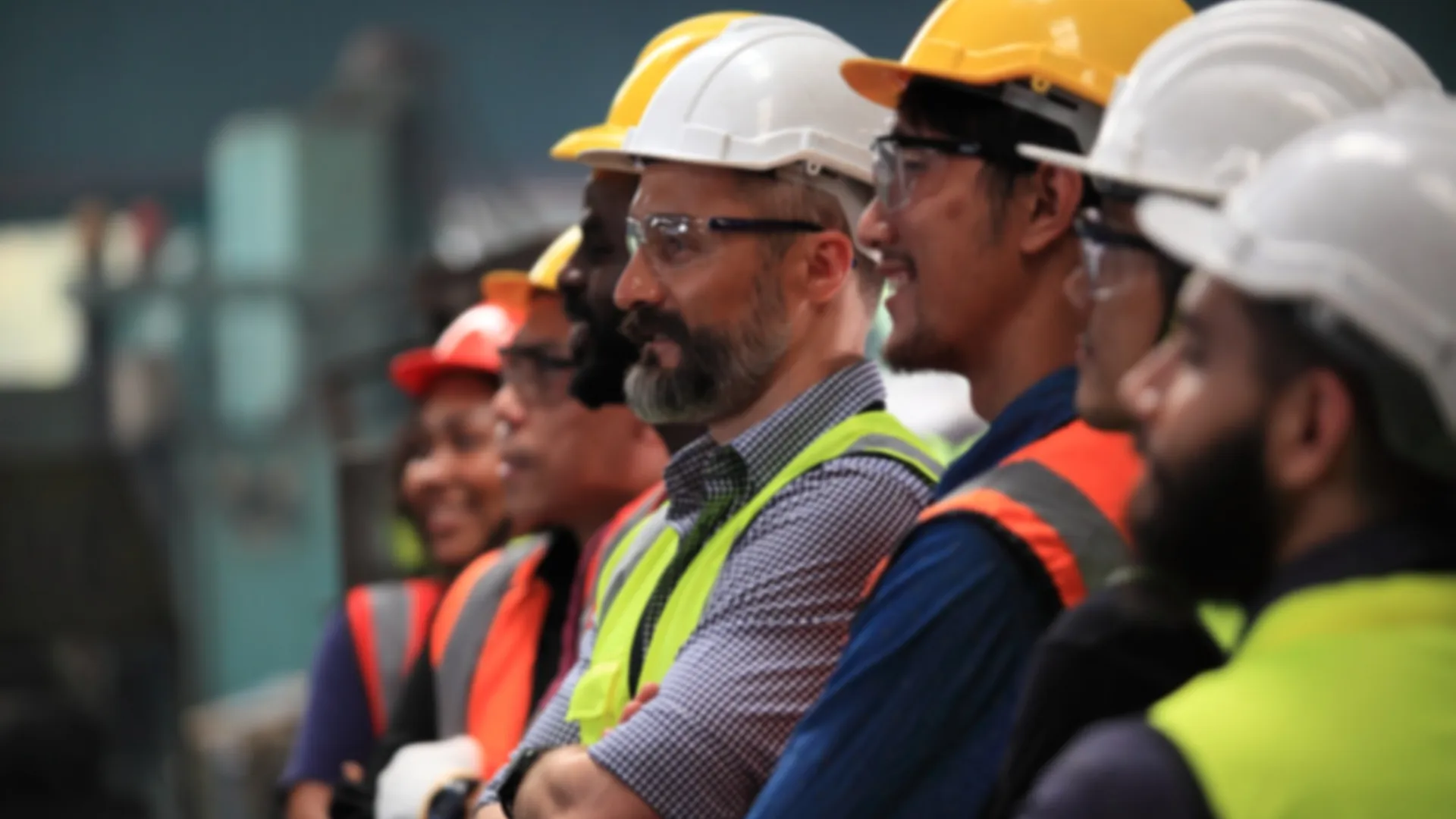Diverse industrial workers wearing safety helmets and vests standing side by side in a factory setting.