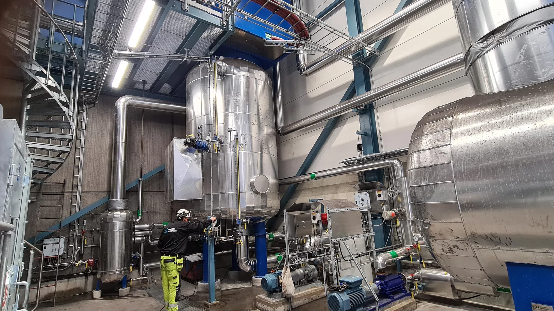 Technician inspecting large stainless steel tanks and industrial equipment in a brightly lit factory setting.