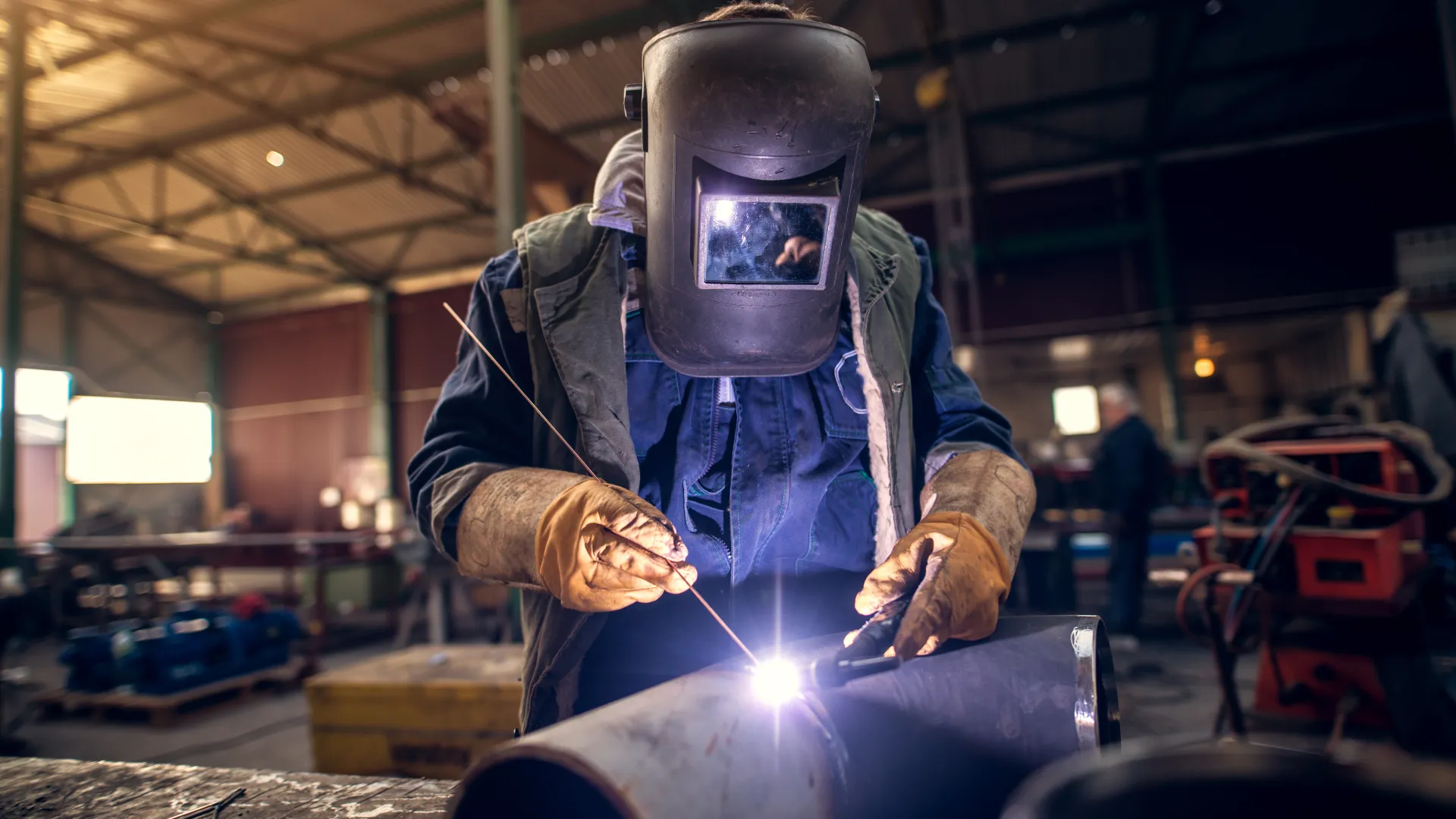 Welder wearing protective gear welding metal pipes in an industrial workshop with sparks flying