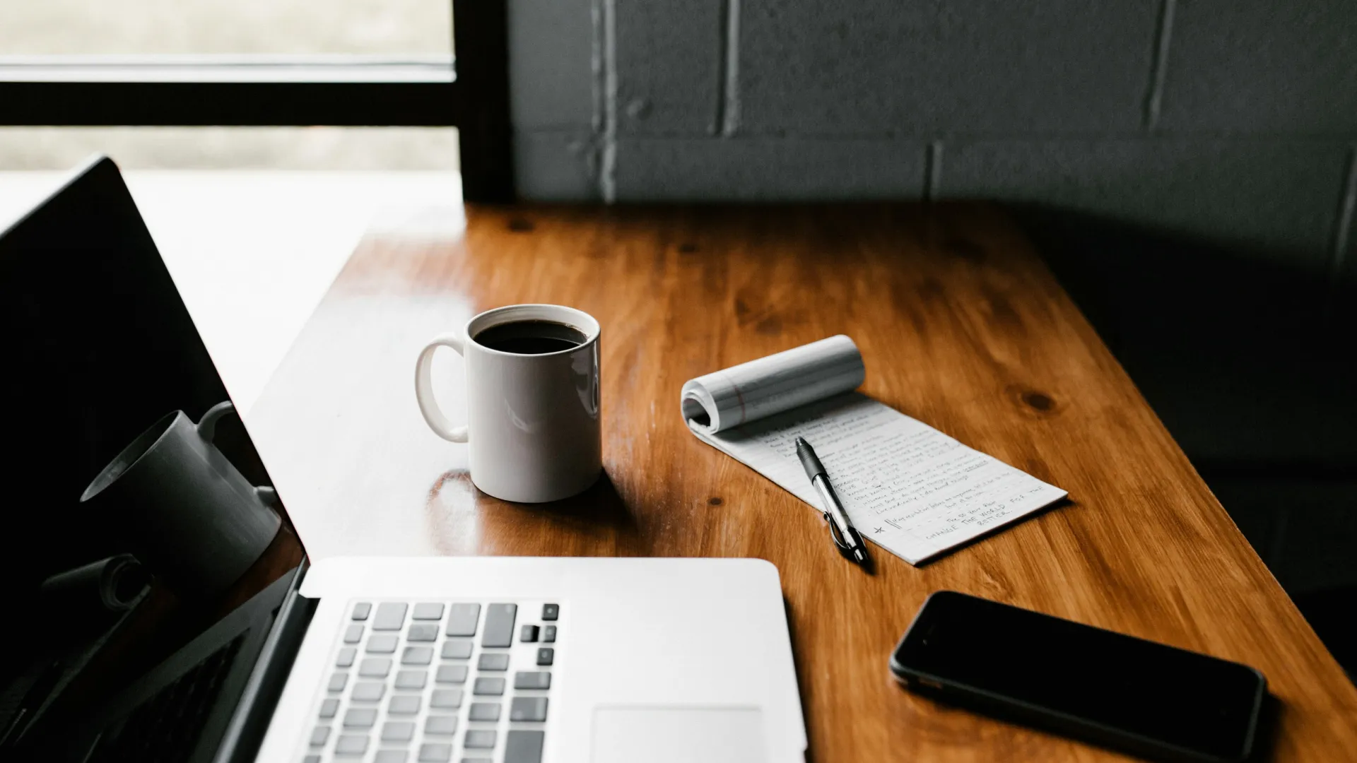 Workspace with open laptop, coffee mug, notepad with pen, and smartphone on wooden table near window.