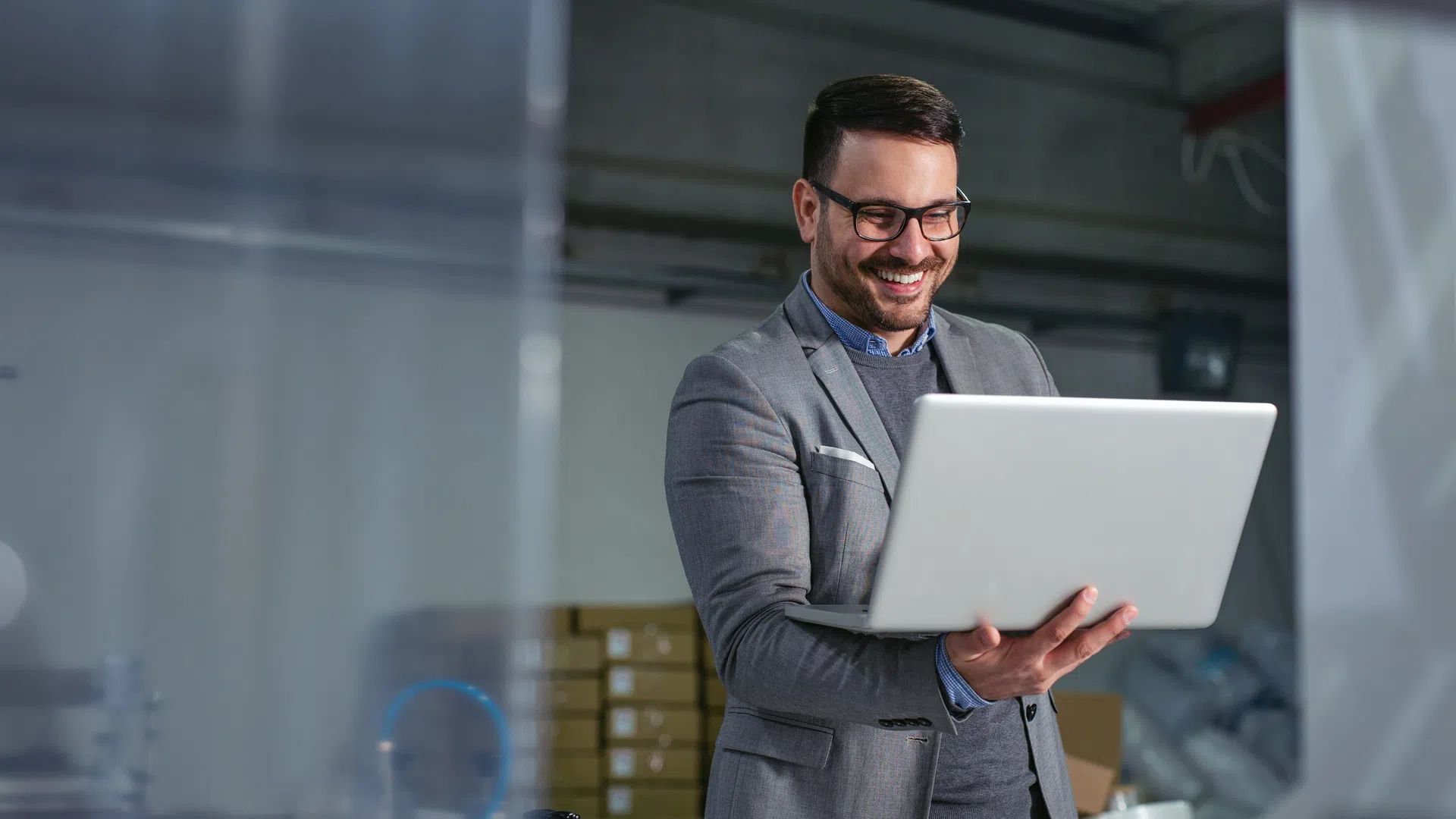 Smiling man in gray suit using laptop in industrial warehouse environment