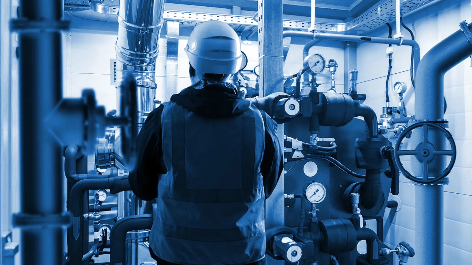 Engineer in safety gear inspecting complex industrial piping and gauges in a mechanical room with blue lighting.
