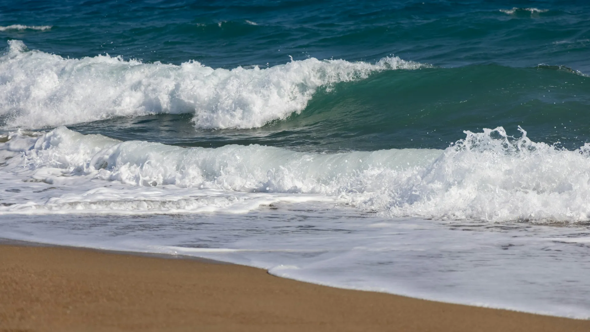 Gentle ocean waves crashing onto a sandy beach under clear blue water and sky.