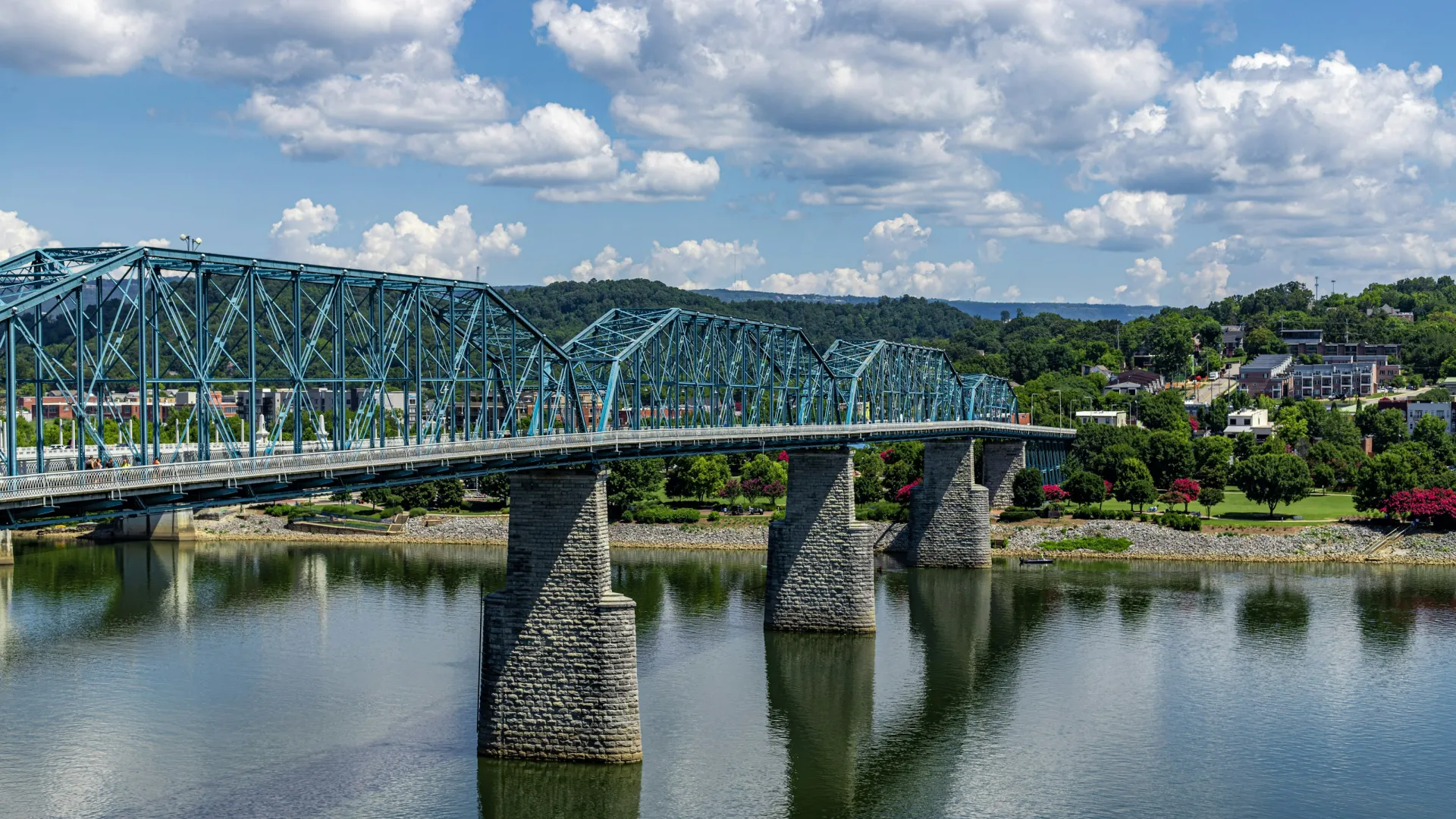 Blue truss bridge spanning a calm river with green landscape and partly cloudy sky in the background