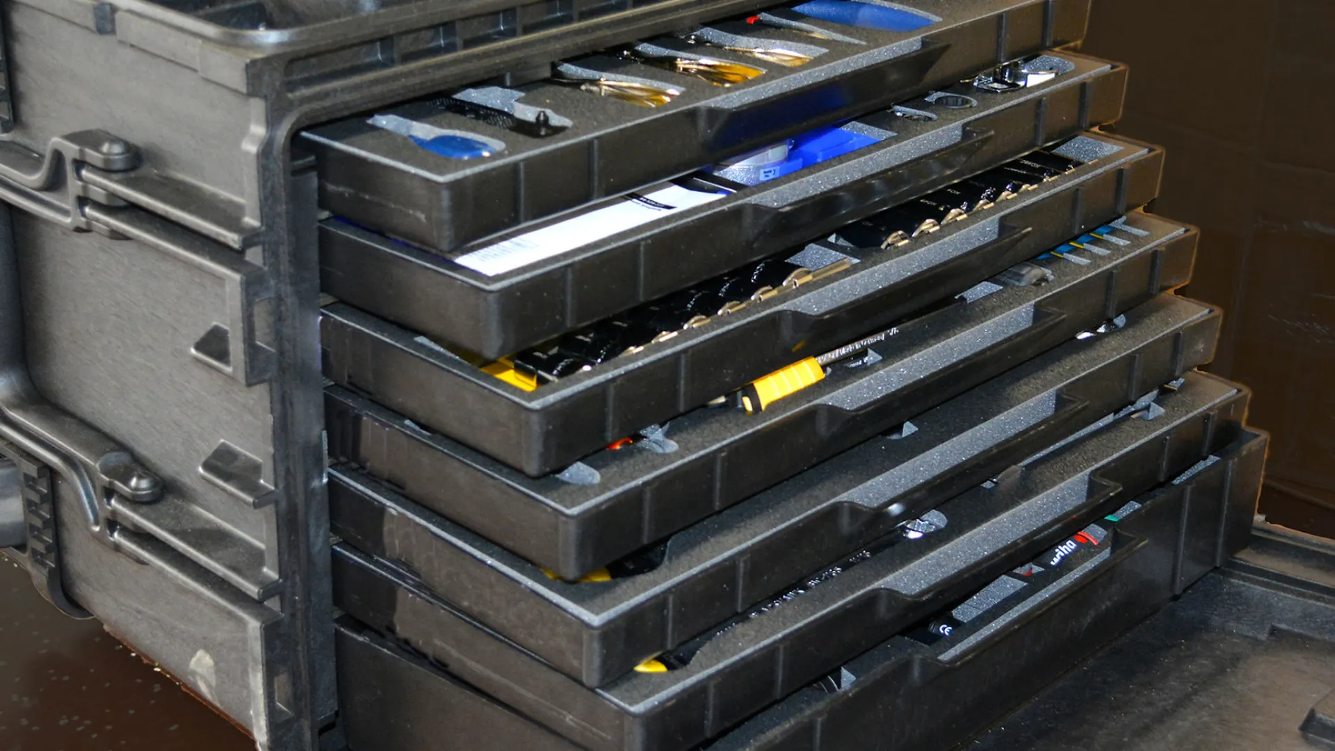 Stack of black plastic trays with tool compartments inside a carrying case, partially open showing organized tools.