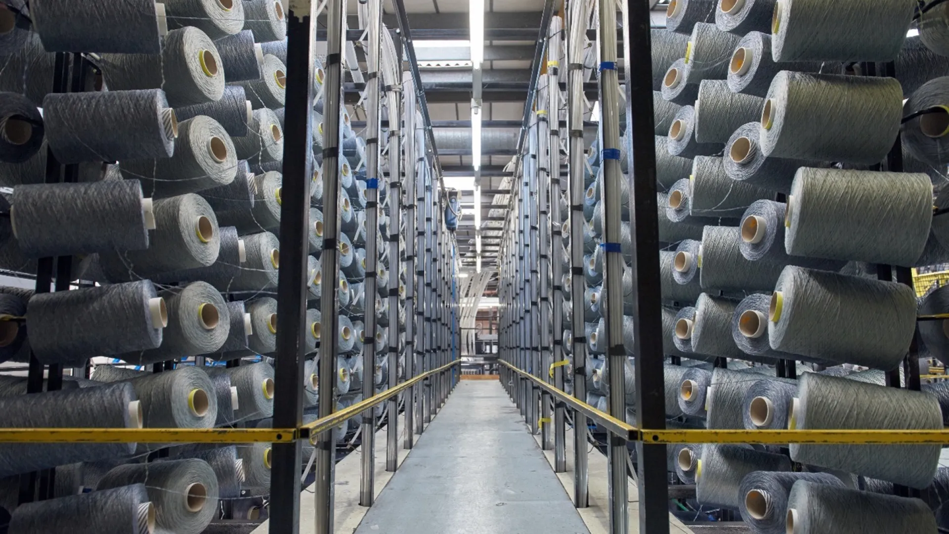 Industrial warehouse aisle lined with large spools of grey textile yarn on both sides under bright overhead lights.