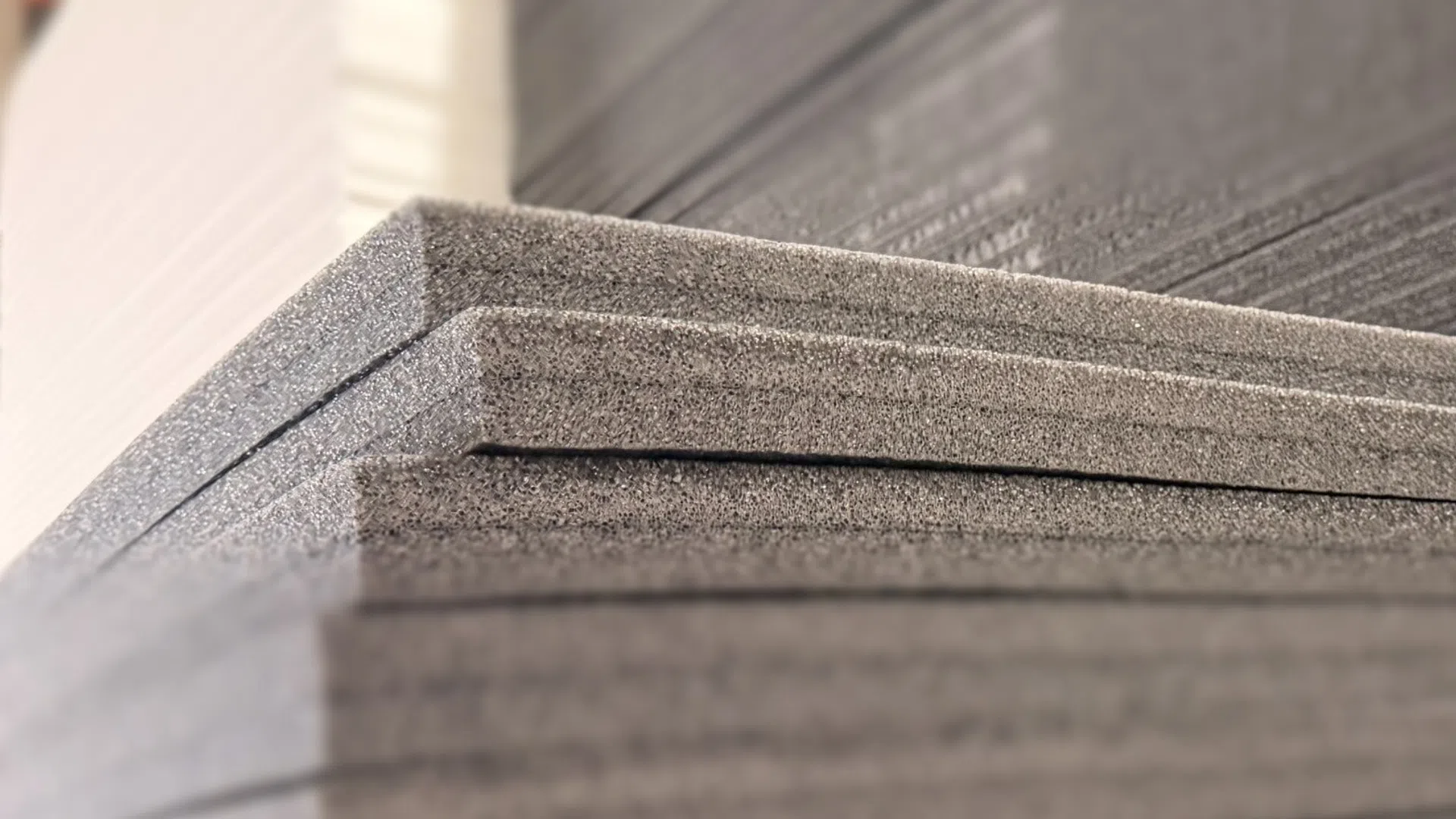 Stack of gray foam sheets in a warehouse against white and black foam panels, showing texture and layers.