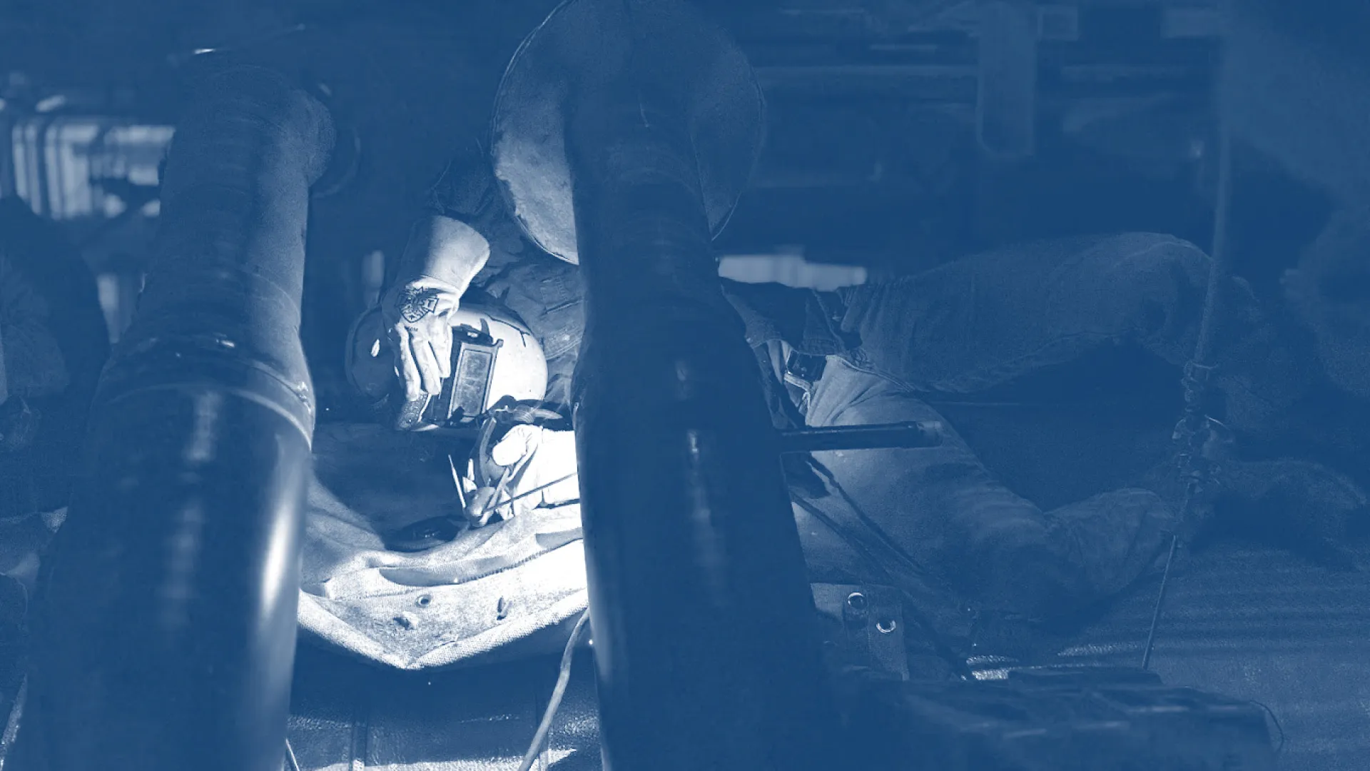 Welder wearing protective gear working under a vehicle with bright welding sparks visible in a dim garage.