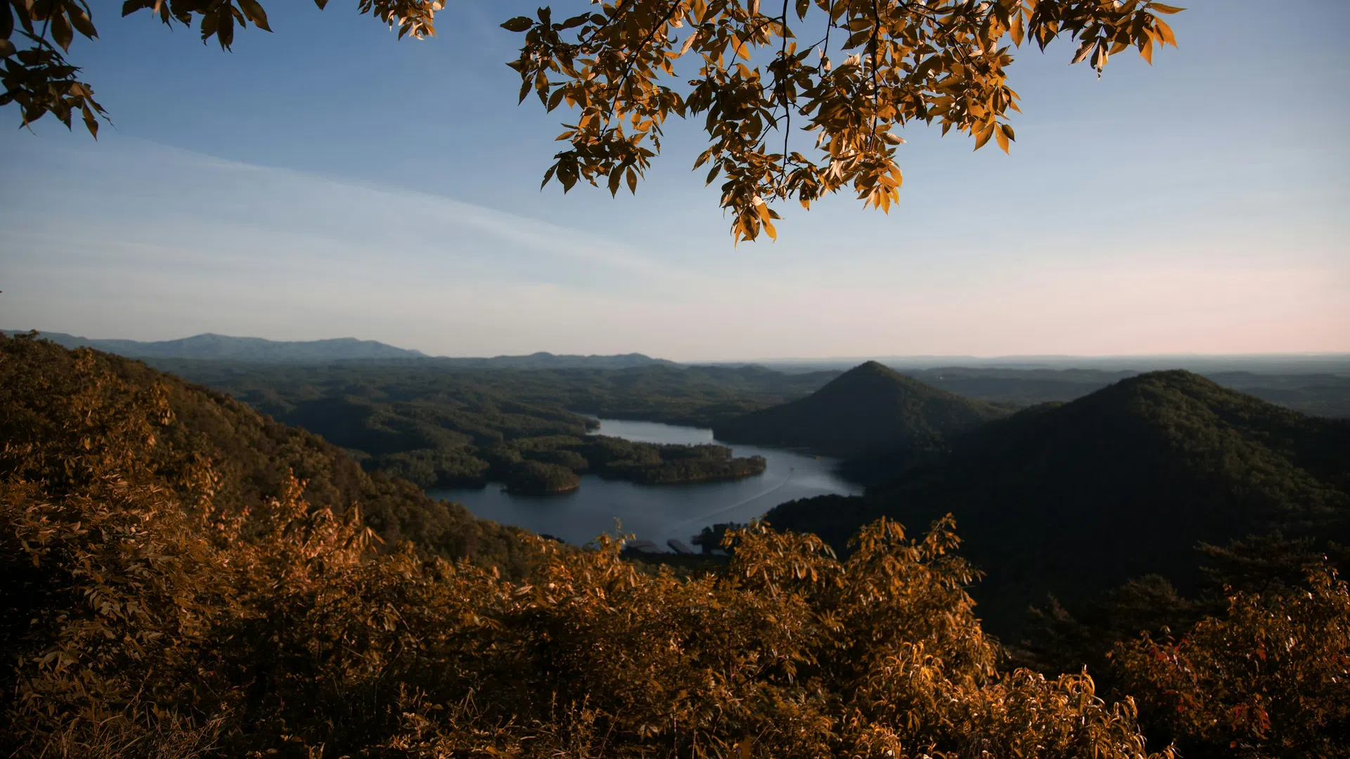 Autumn leaves frame a scenic lake and mountain landscape under a clear blue sky at sunset.