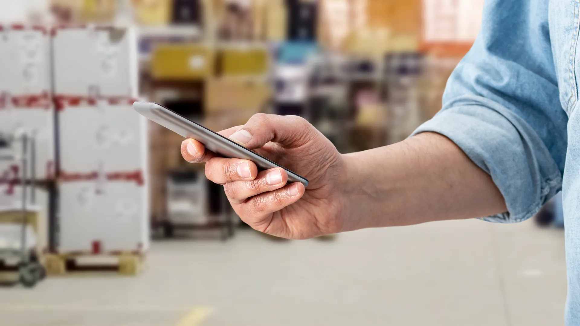 Person using smartphone in a warehouse with shelves and boxes in the background, wearing a rolled-up denim shirt.