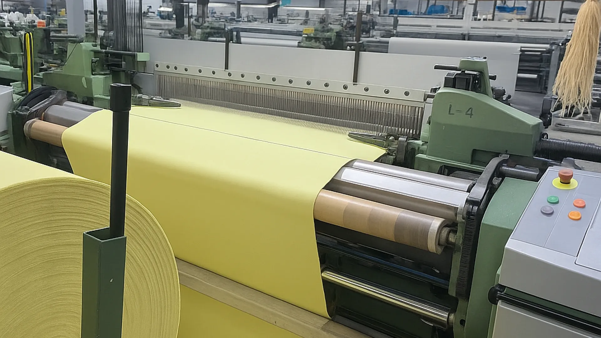 Industrial weaving machine producing wide rolls of yellow fabric in a spacious textile factory.