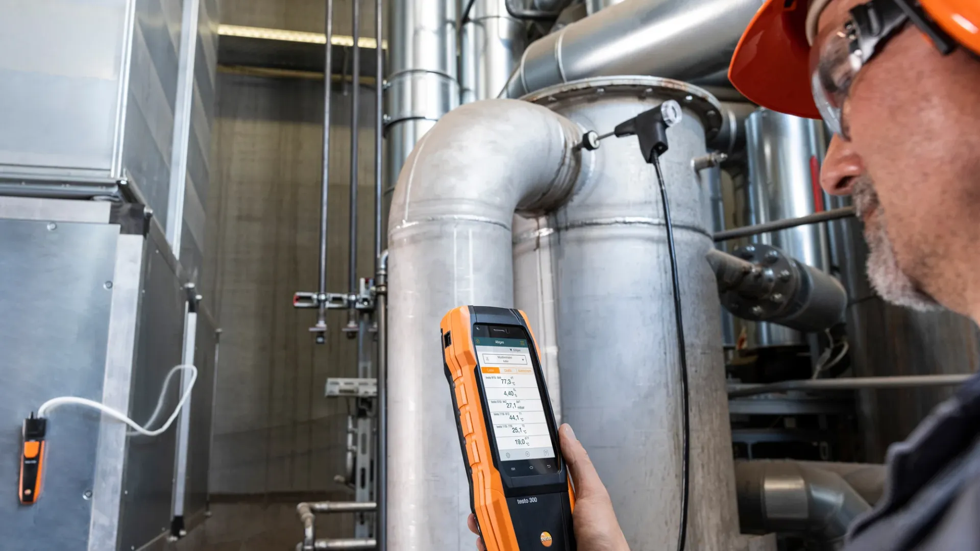 Technician using an orange handheld device to inspect metal industrial pipes and valves in a factory setting.