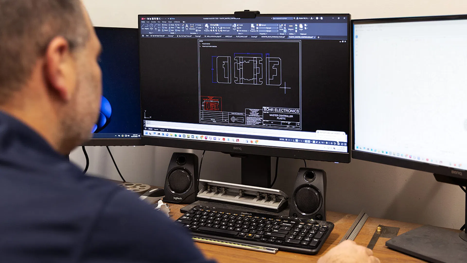 Engineer designing electronic component schematics on a computer with three monitors and speakers in an office.