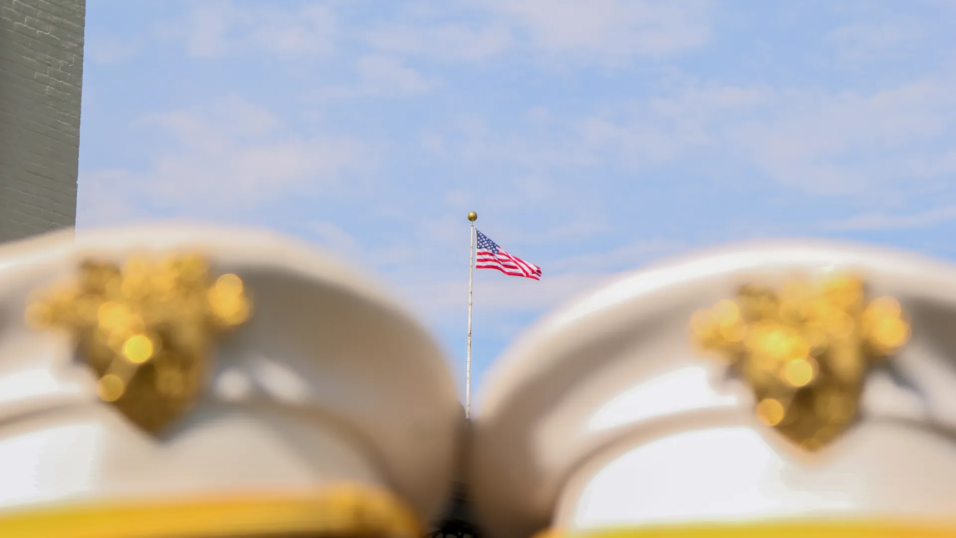 American flag waving in the sky framed between two blurred white military hats with gold insignia.