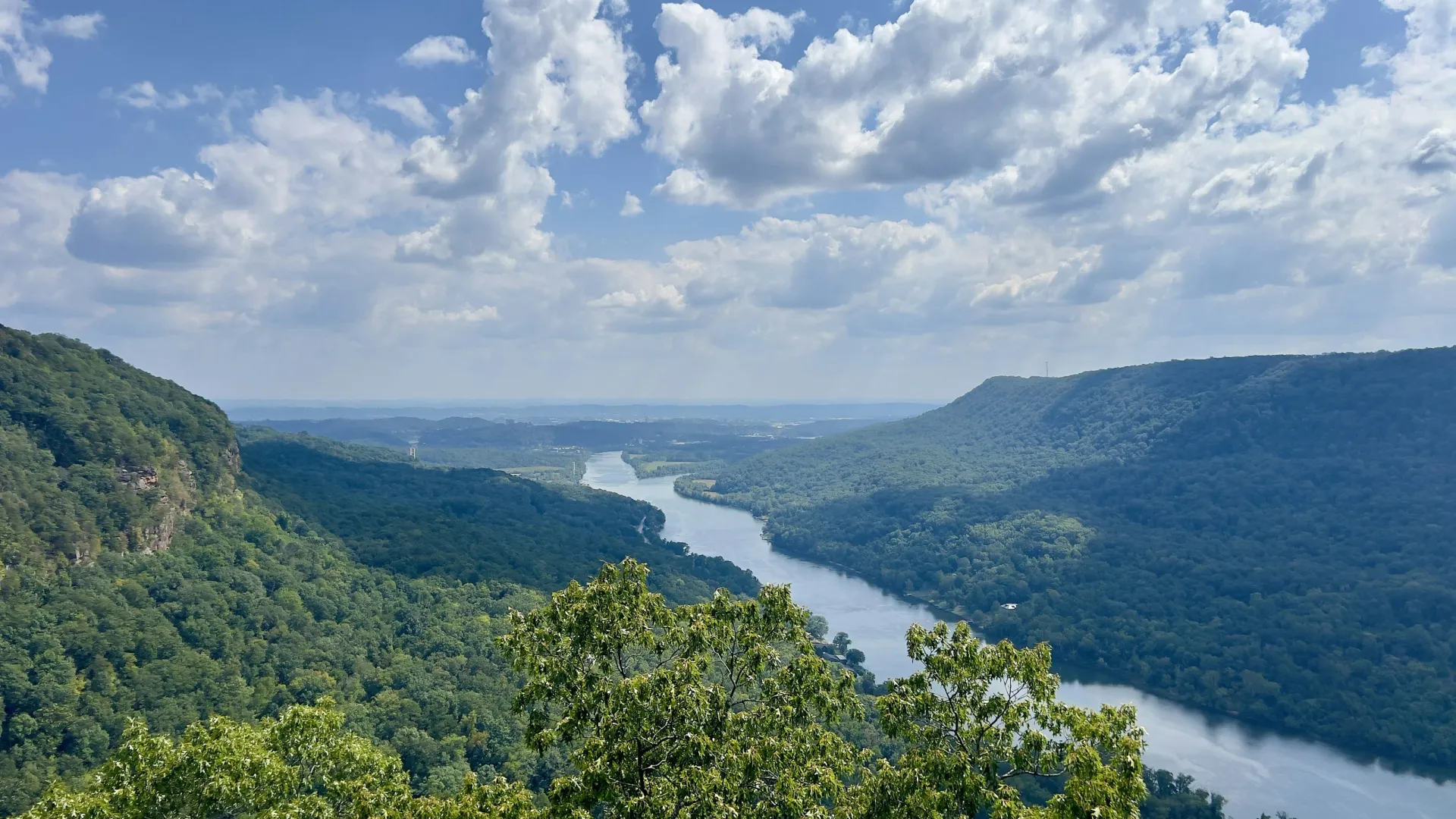 Wide river flowing through lush green forested mountains under a blue sky with scattered clouds