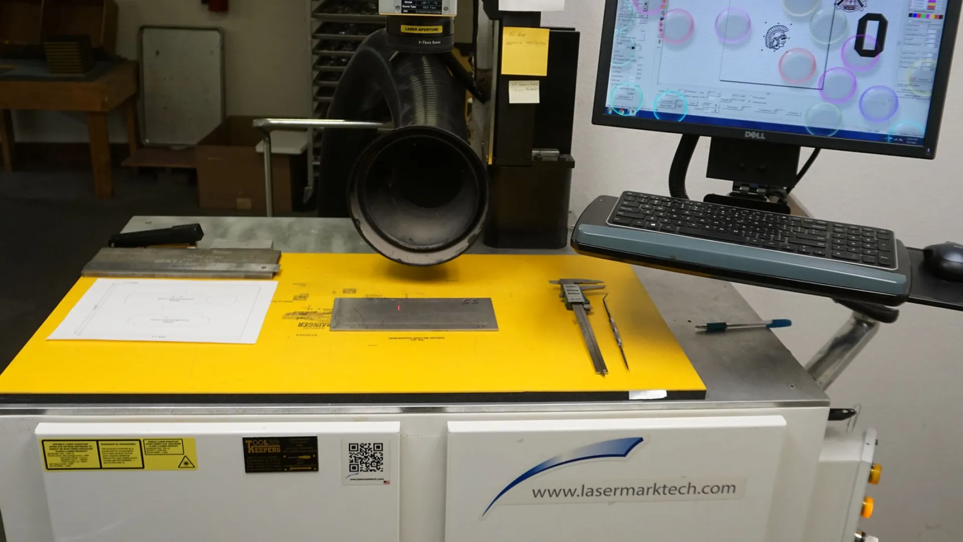 Industrial laser marking machine with computer control and measuring tools on a yellow work surface in workshop.