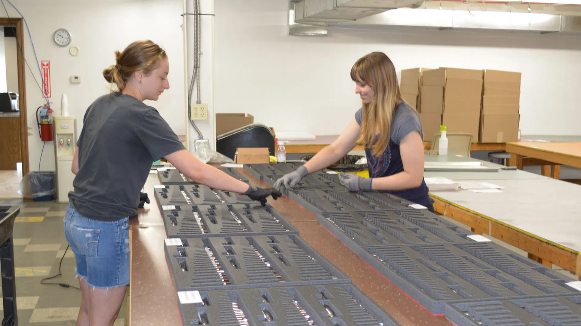 Two women assembling or inspecting organized trays of small items on a large industrial table in a warehouse.