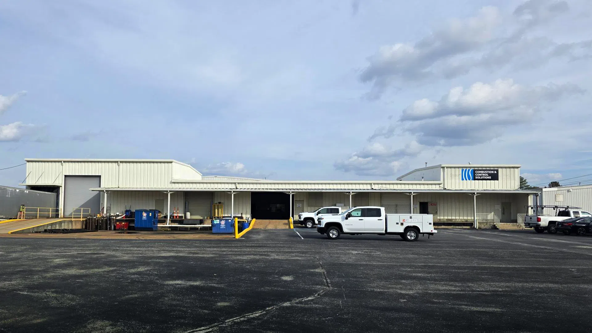 Industrial warehouse building with trucks parked outside under a cloudy sky on a large asphalt lot.