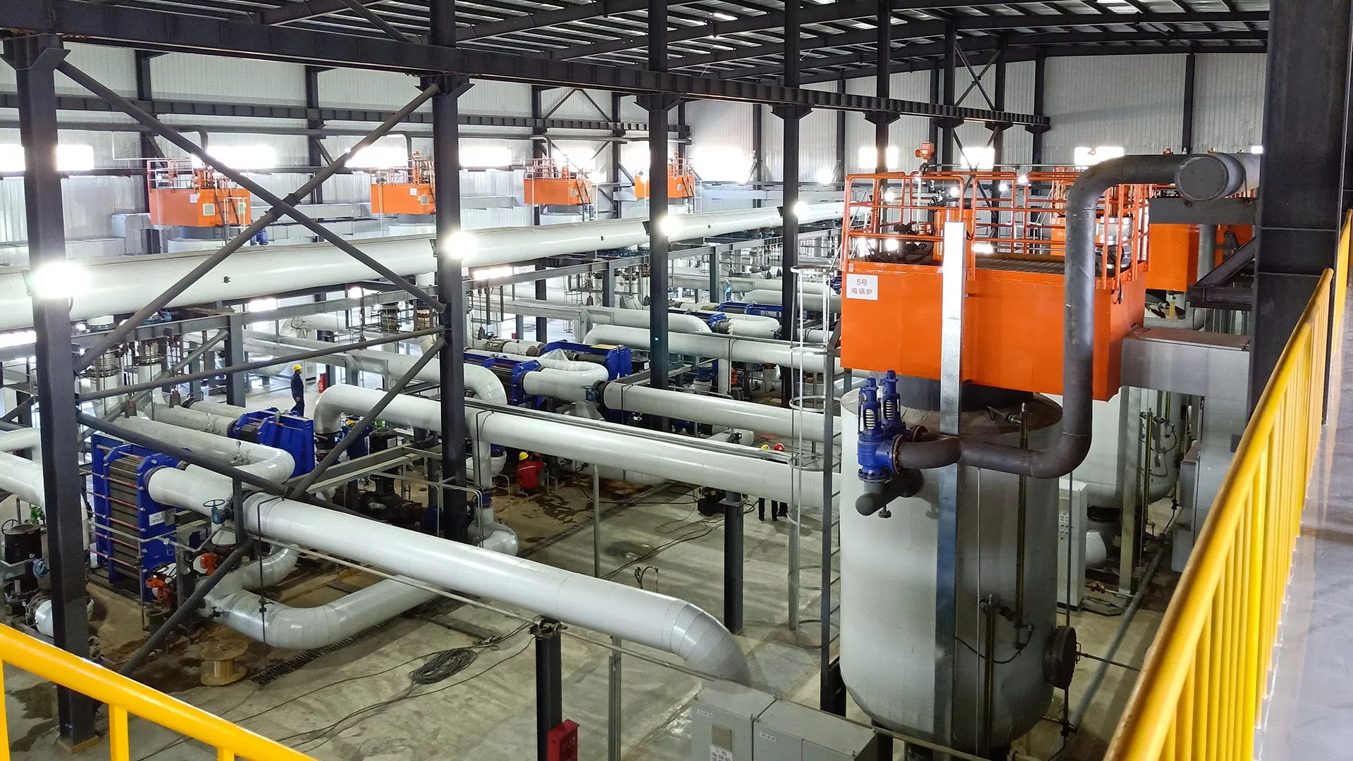 Industrial facility interior with large white pipes, orange tanks, and black steel framework under a metal roof.