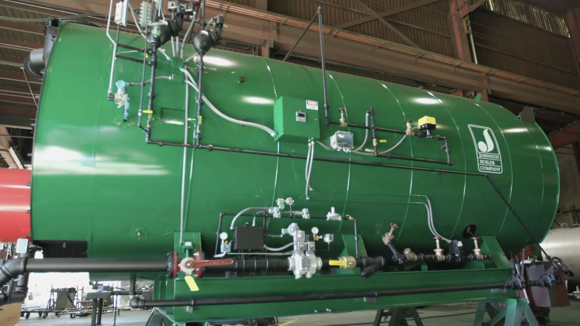 Large green industrial boiler with pipes and gauges inside a factory setting under metal roofing.