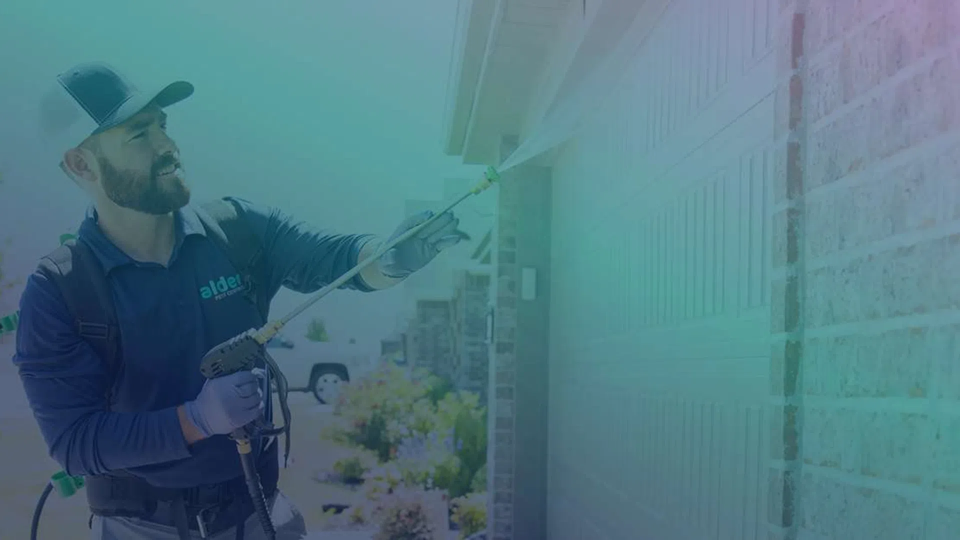 Pest control technician spraying insecticide on home exterior near garage door in bright daylight.