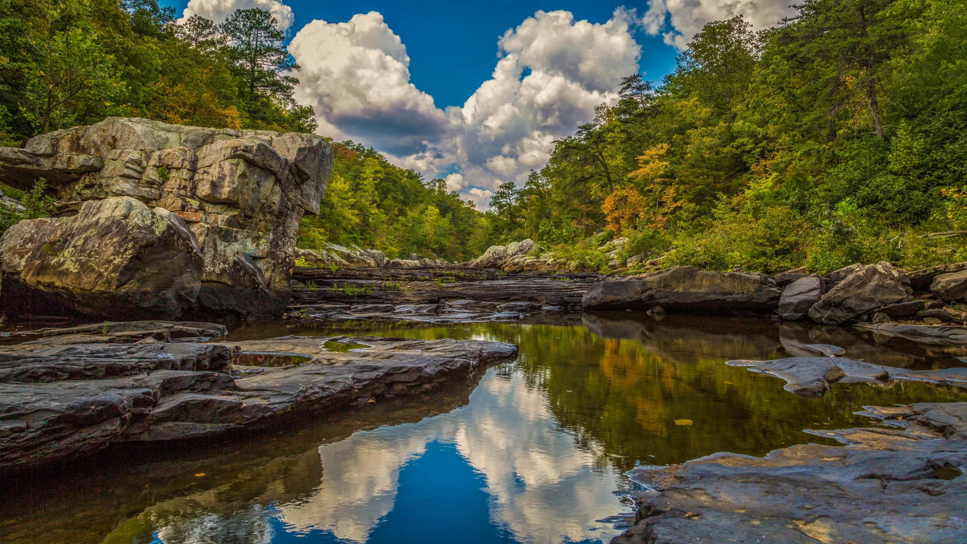 Tranquil river landscape with rocky formations and vibrant autumn foliage under a blue sky with clouds.