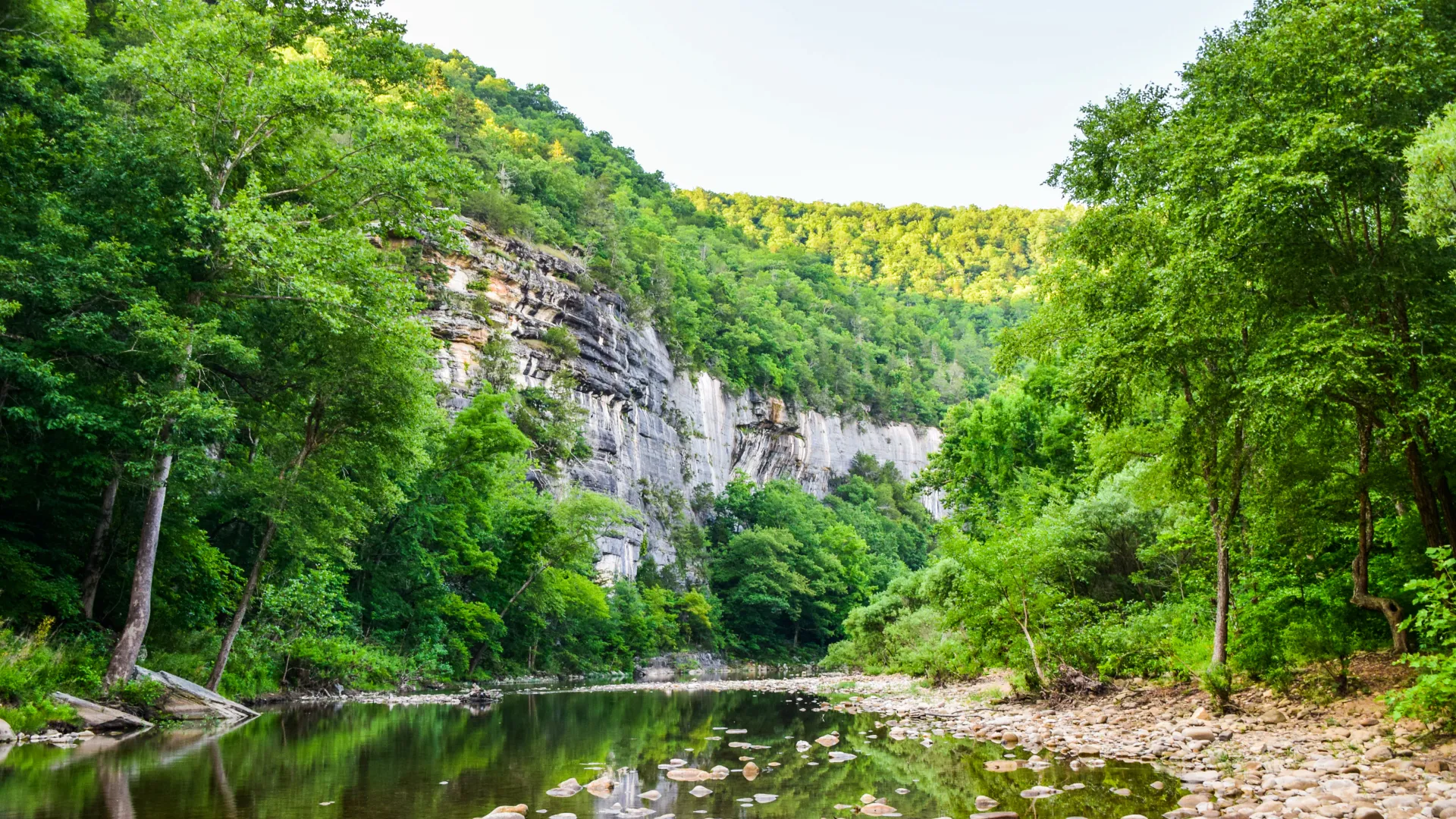 Serene river flowing through lush green forest and towering cliffs under clear blue sky.