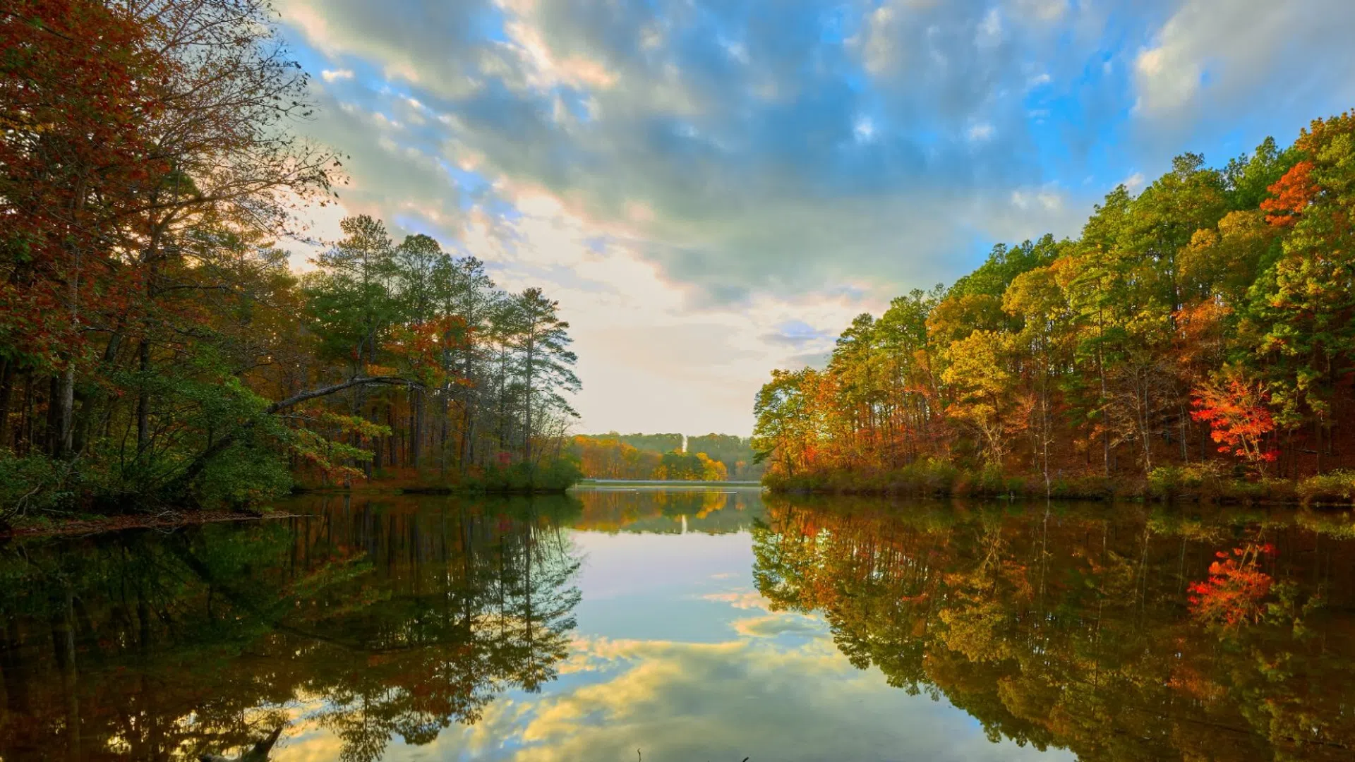 Autumn trees with colorful foliage reflected in the calm river under a partly cloudy sky.