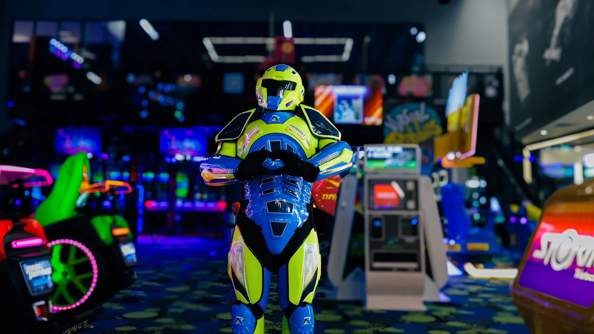 Person wearing neon green and blue armored suit standing in a dimly lit arcade with colorful game machines.