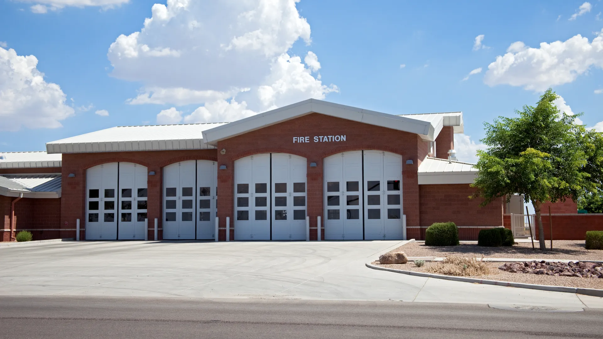 Red brick fire station with five white garage doors under a bright blue sky with fluffy clouds.