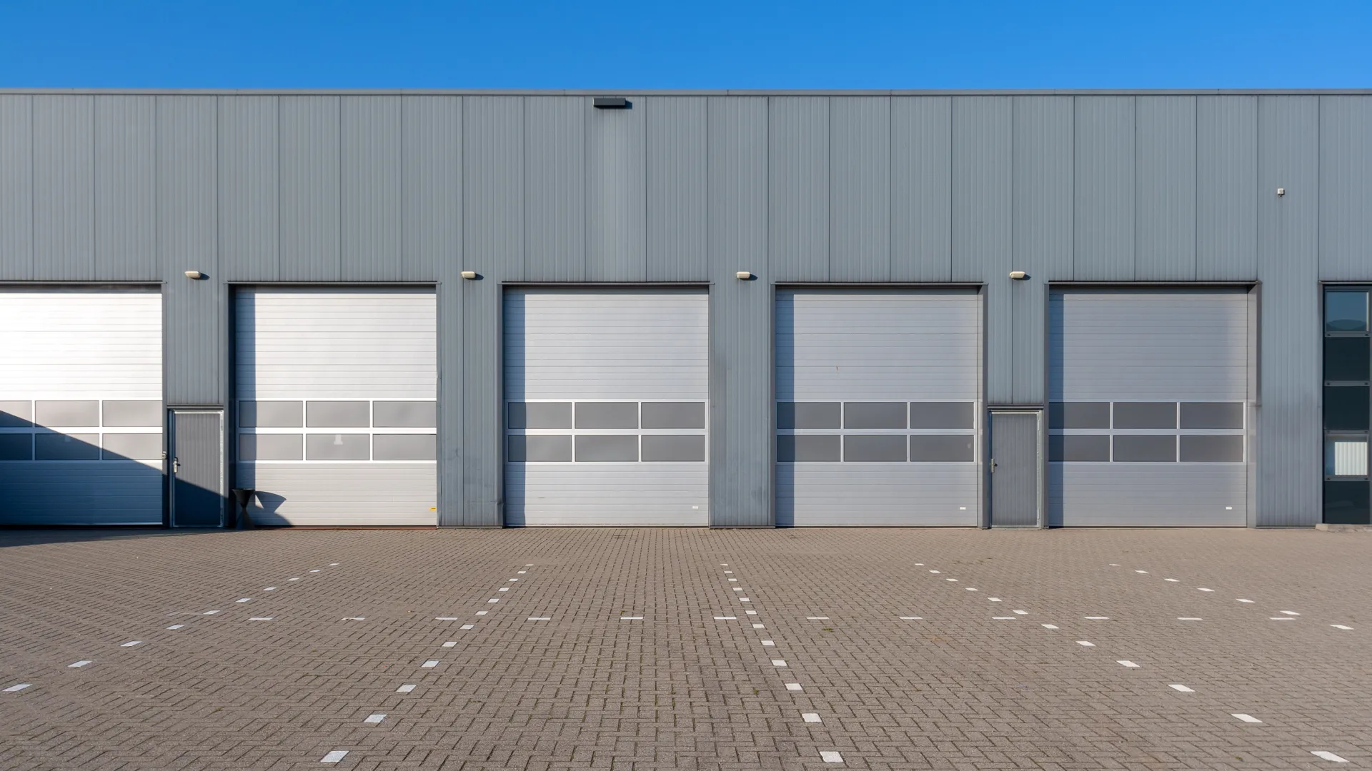 Empty industrial warehouse exterior with five closed gray loading dock doors and a paved parking area under clear blue sky.