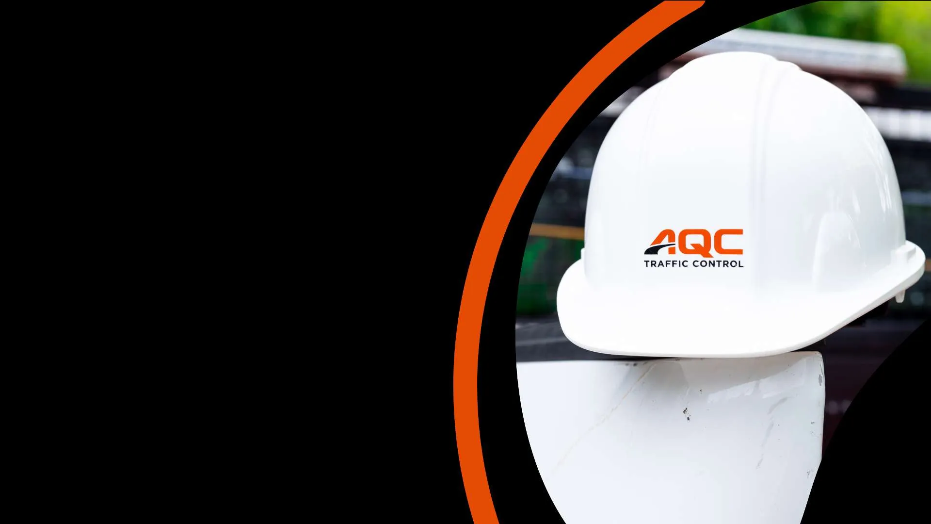 White construction hard hat with AQC Traffic Control logo resting on a white barrier with black and orange background.