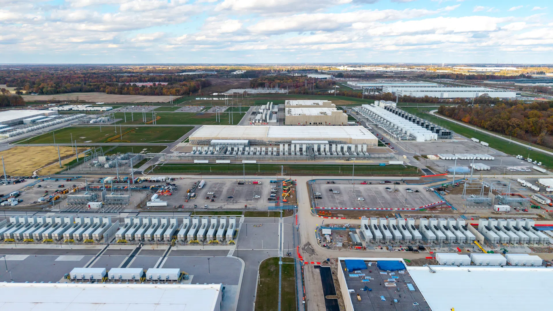 Aerial view of a large industrial data center complex with multiple buildings, parking lots, and trailers under a cloudy sky.