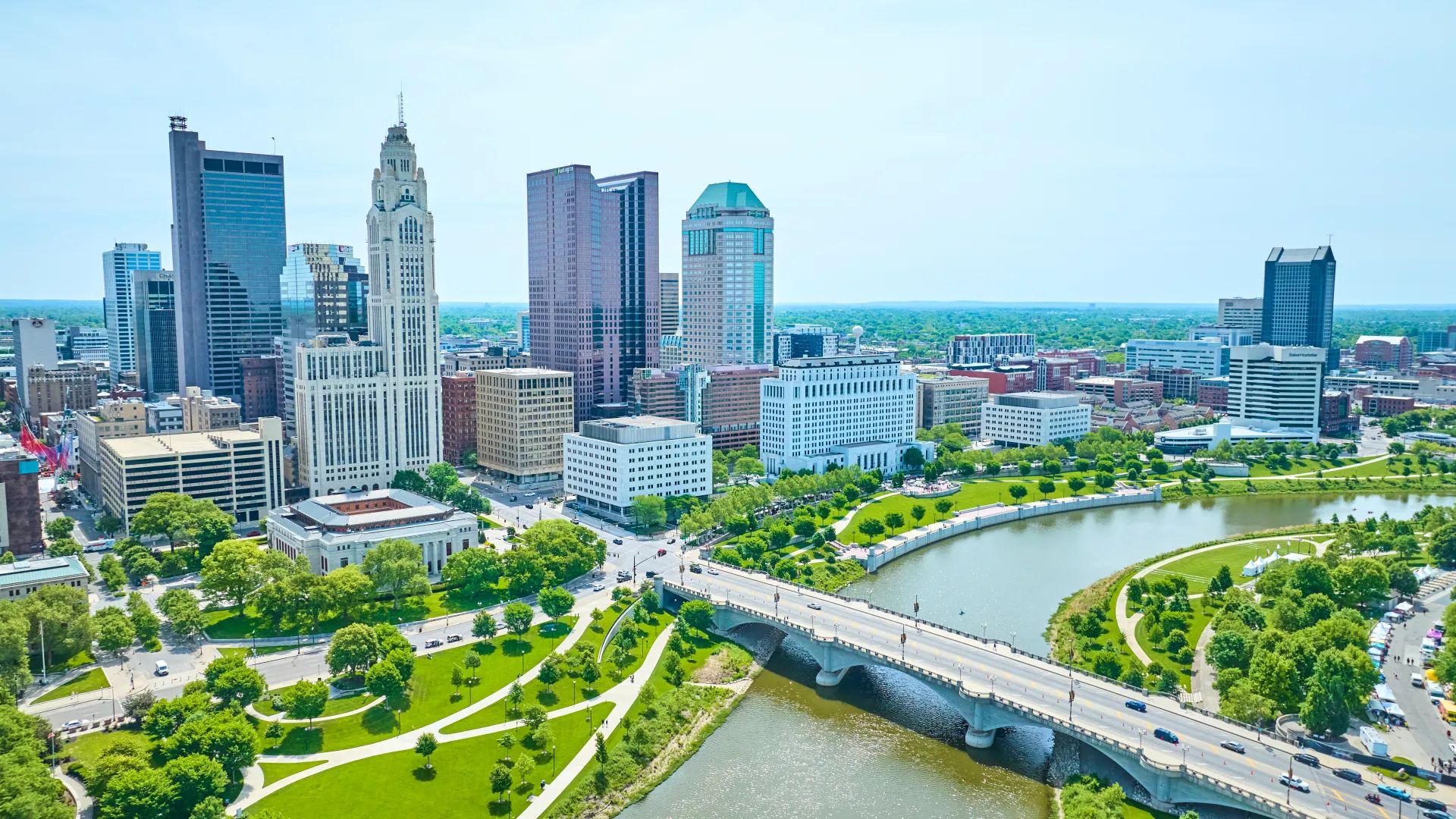 Aerial view of Columbus Ohio skyline with river, bridge, and green park space on a clear sunny day