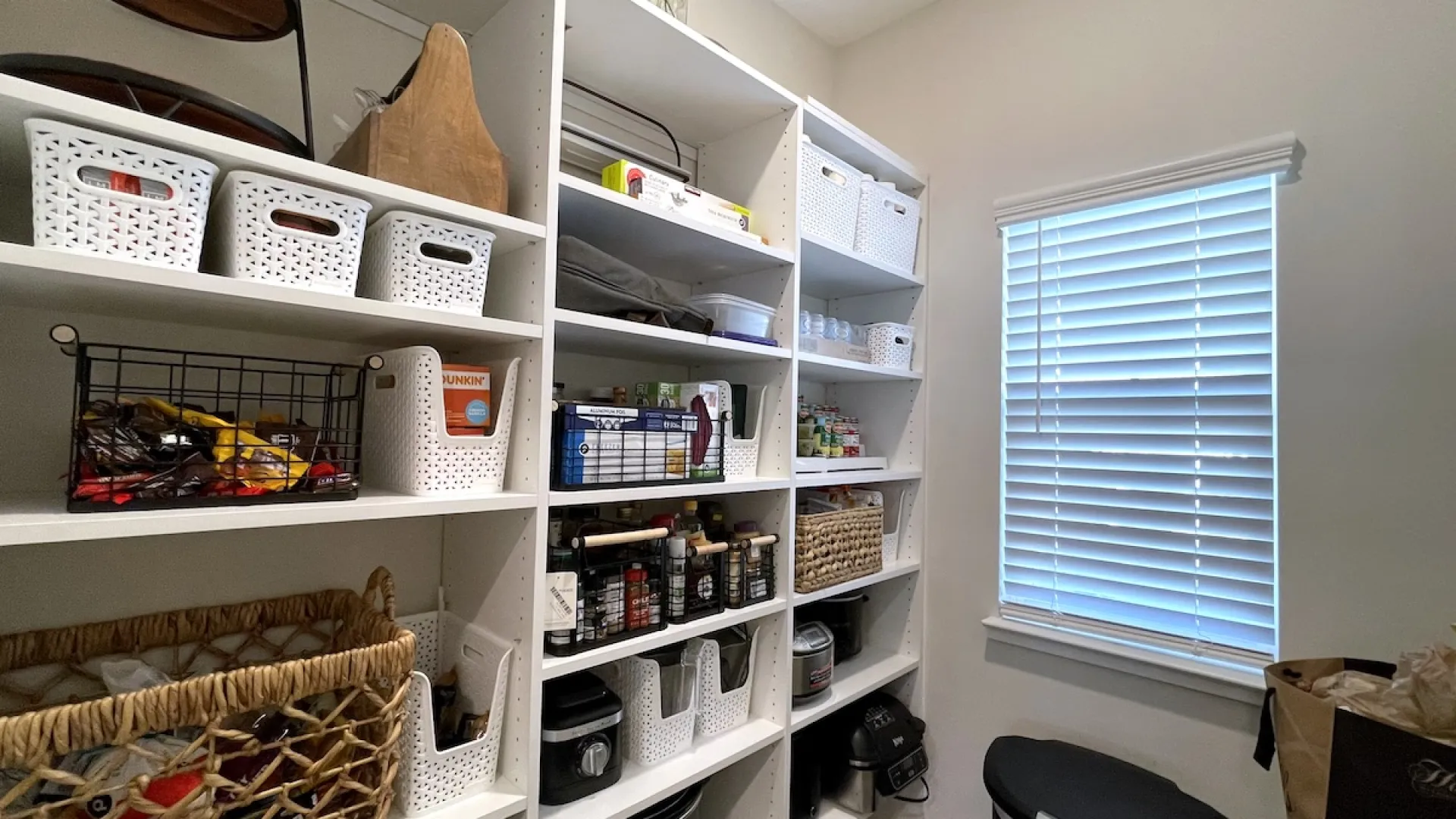 Organized pantry shelves with white baskets, containers, spices, and kitchen items next to a window with white blinds.