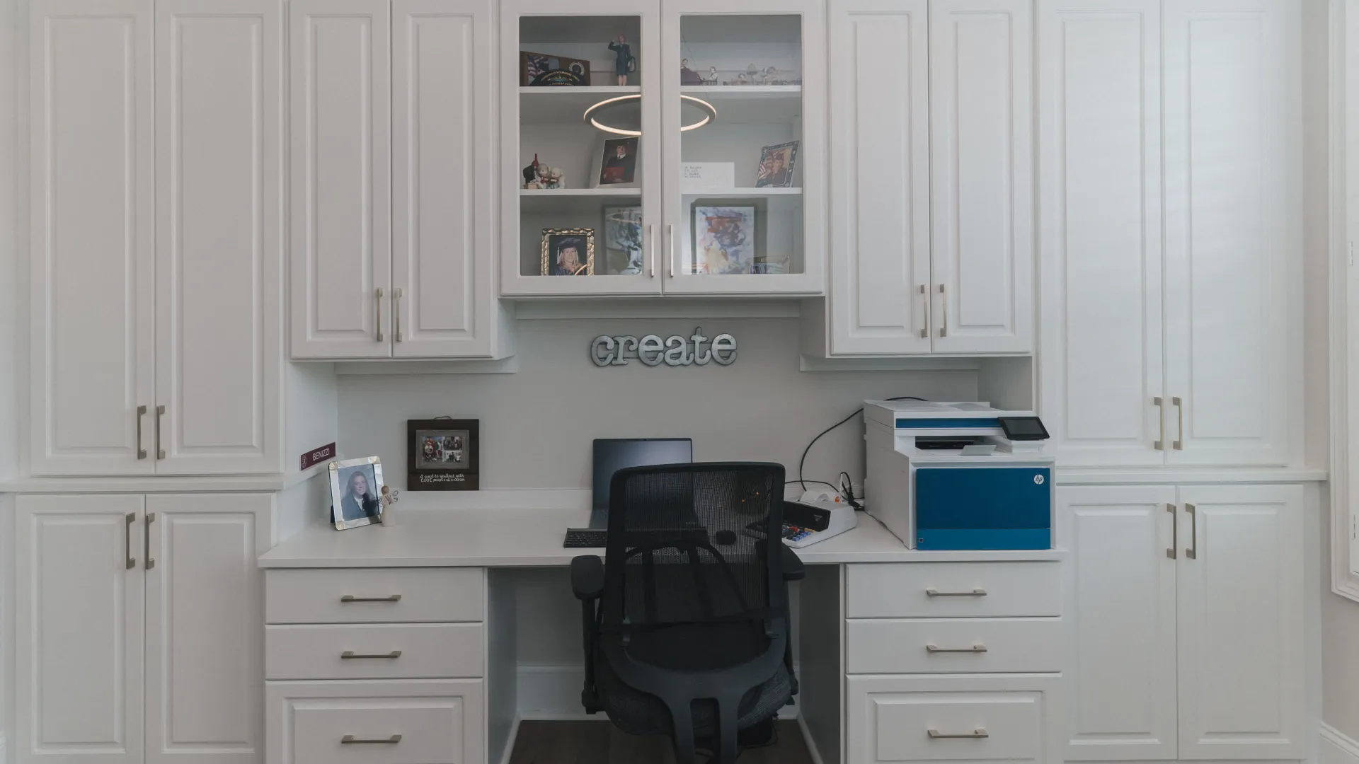 modern home office with white desk drawers, white cabinets, glass cabinet doors, laptop, printer, black executive chair and decorative create sign on wall.