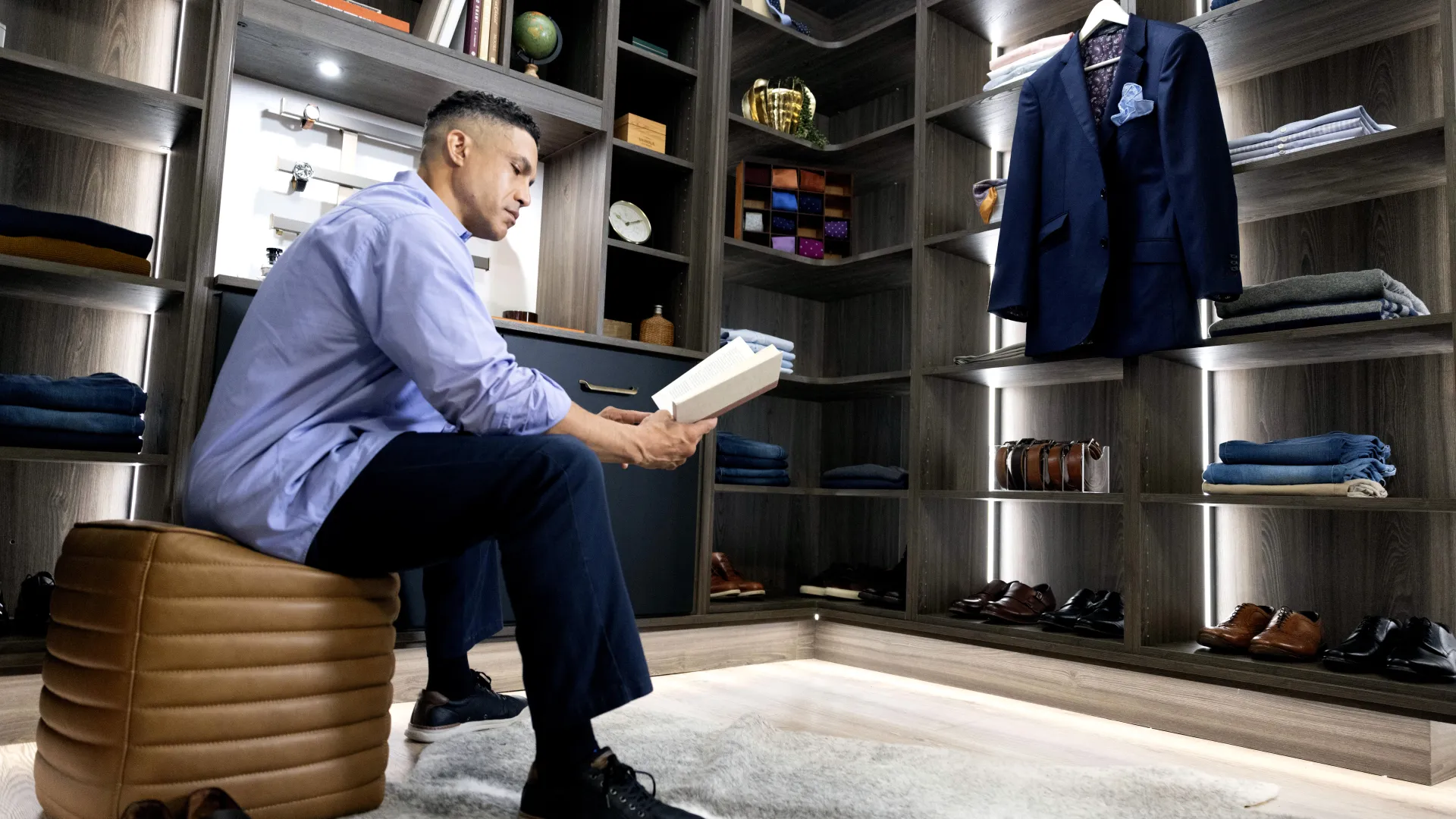 Man sitting on a ottoman reading a book inside a modern walk-in closet with clothes and shoes.