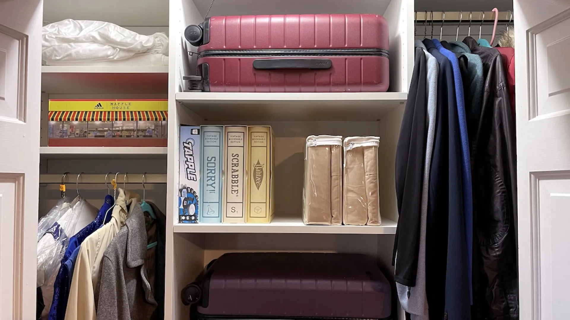 Organized closet with hanging clothes, two suitcases, board games, and folded items on shelves inside white closet.