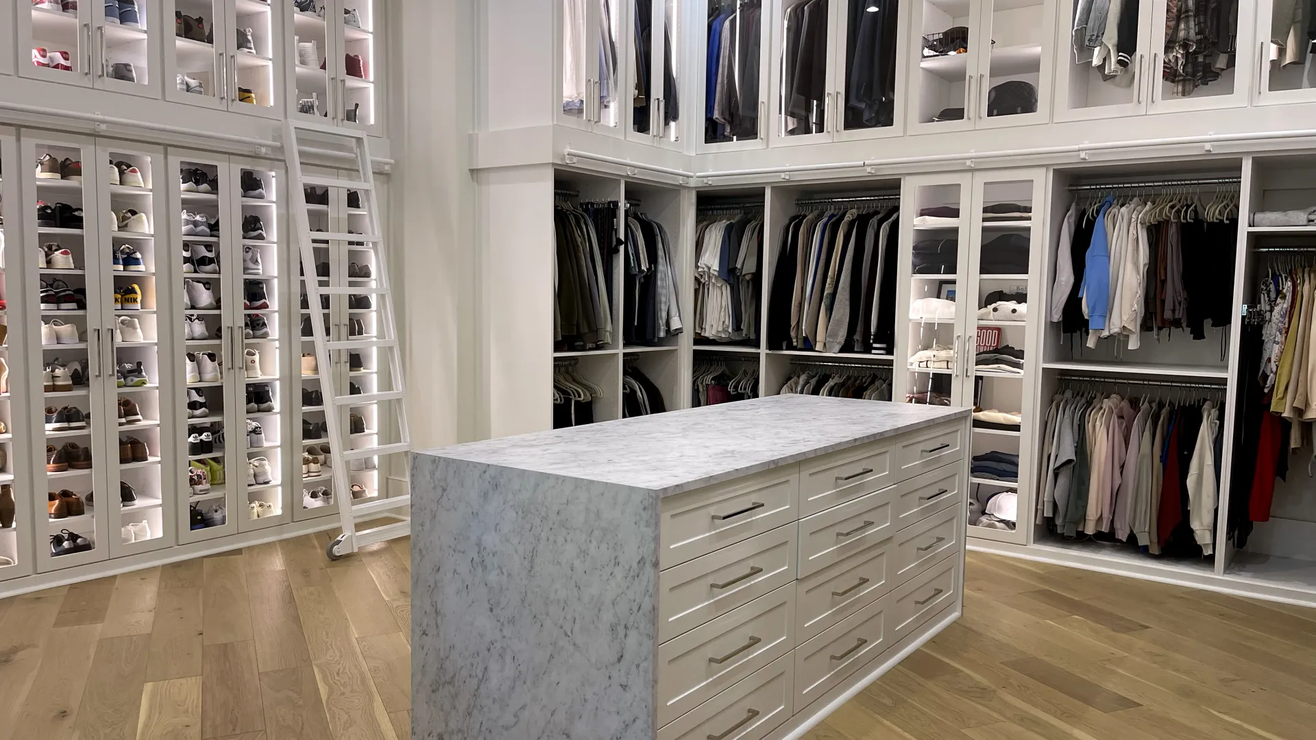Custom walk in closet with lighting, white cabinetry, marble island, shoe shelving with glass doors, mens clothing, womens clothing, shoes and a white ladder