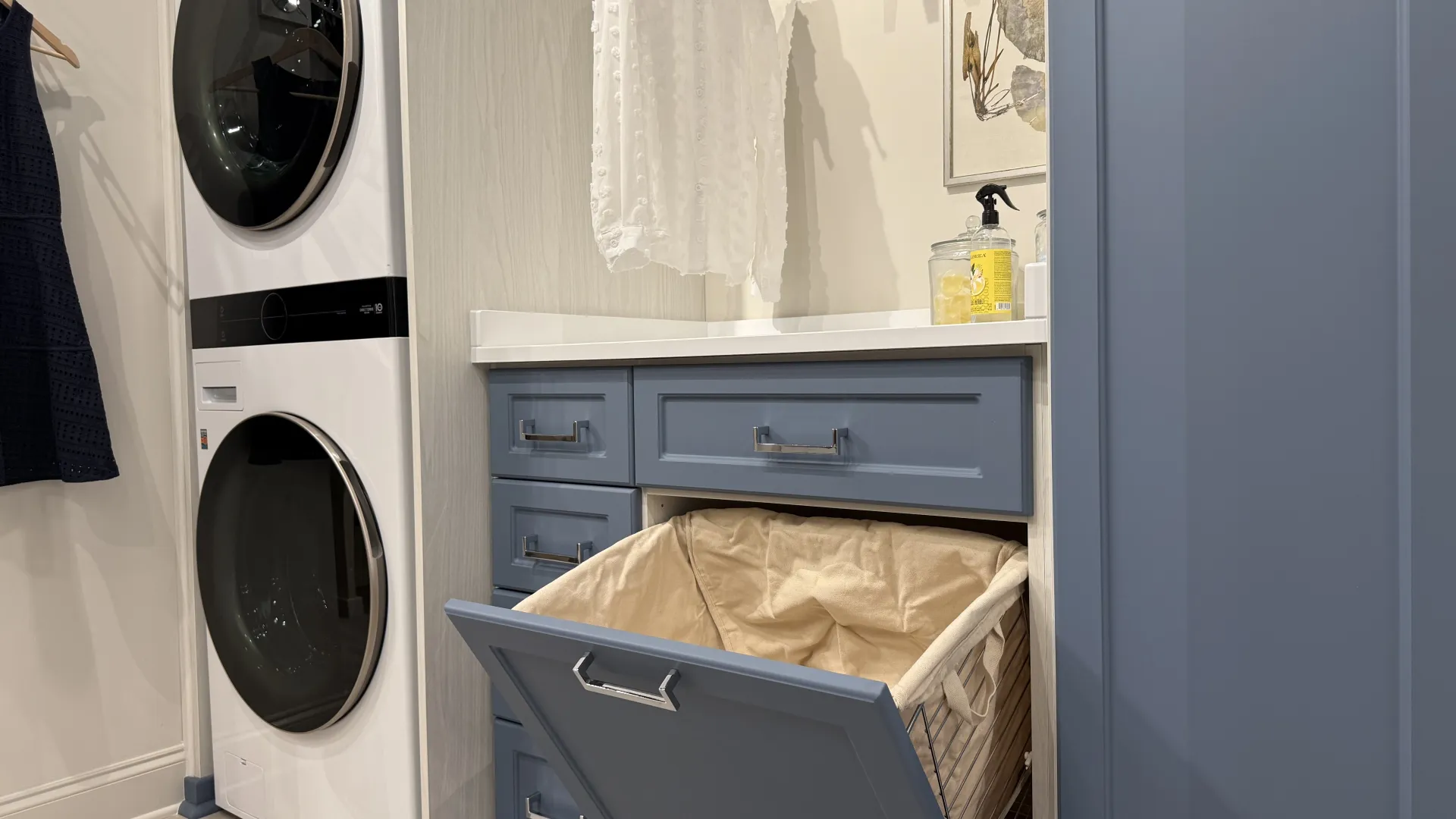 Modern laundry room with stacked washer and dryer, blue cabinets, and built-in flip hamper with beige liner.